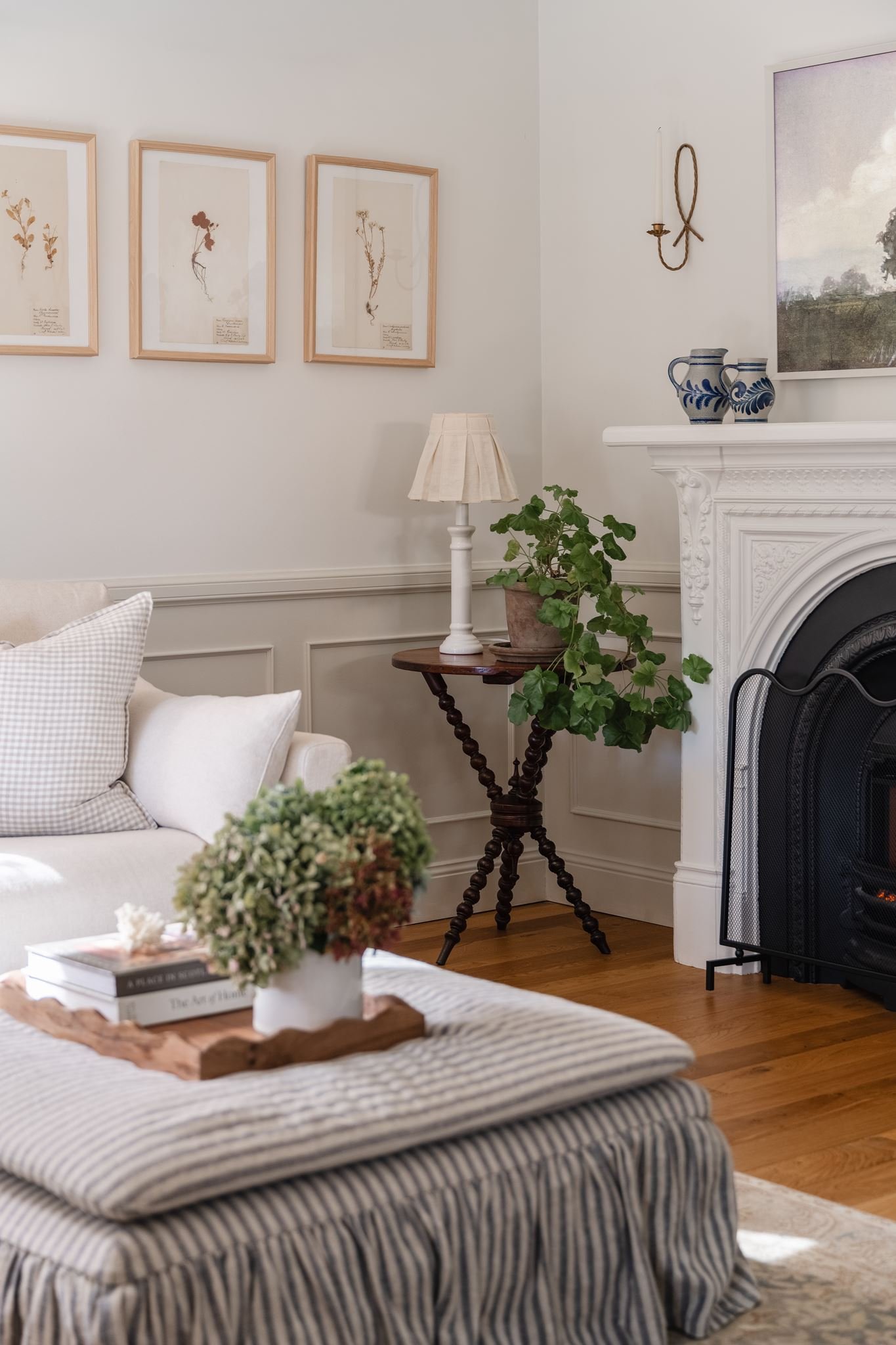 A cozy living room with a white sofa, striped ottoman, and a coffee table with books and a floral arrangement. There is a white fireplace mantel with blue and white vases, a framed landscape picture, a wall-mounted candle holder with a rope design, a