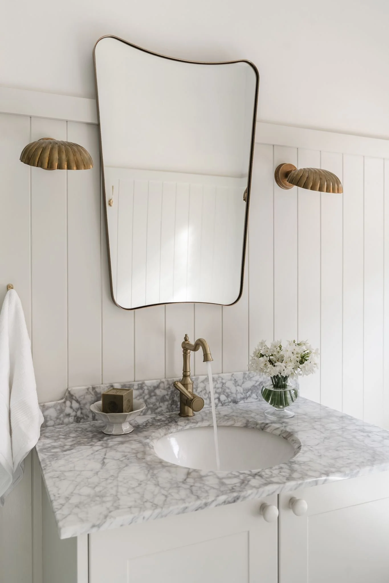 Bathroom sink with white cabinetry, marble countertop, brass faucet, oval mirror, brass wall sconces, a white towel, a small soap dish, and a vase with white flowers.
