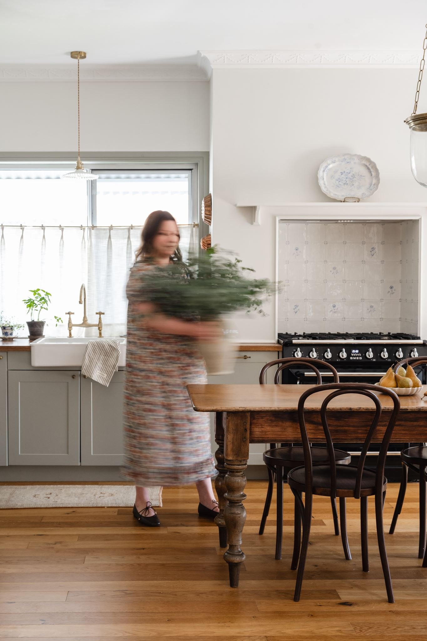 A woman with long dark hair wearing a patterned dress and black shoes is walking through a kitchen, slightly blurred due to motion. The kitchen has a wooden table, black chairs, and a stove. There are white cabinets, a white sink with a gold faucet, and a window with sheer curtains. Potted plants sit on the counter.