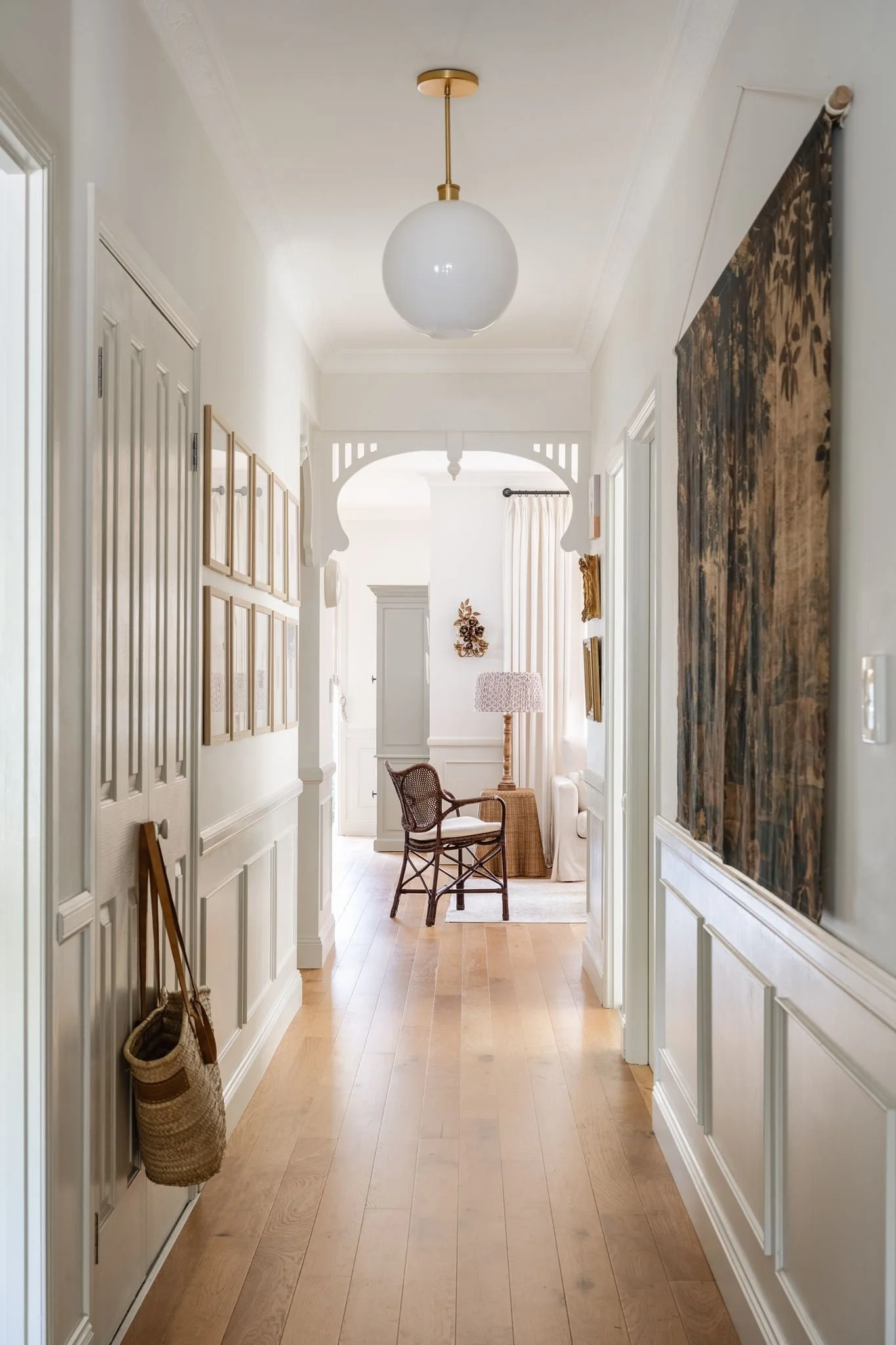 A bright hallway with wooden flooring, white walls, and decorative molding. On the left, there are closed closet doors and a woven basket hanging on a hook. On the right, a tapestry hangs on the wall. Light from the window at the end illuminates a co