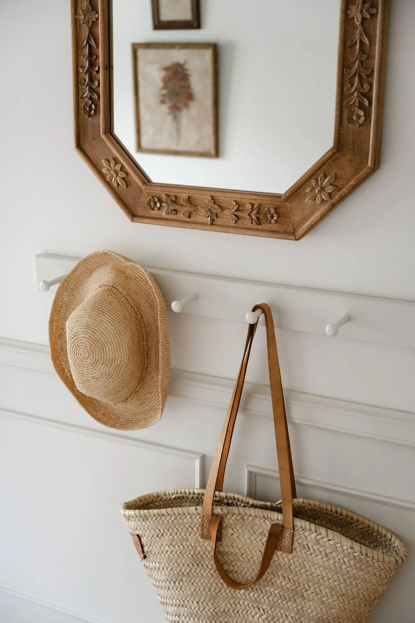 A white wall with a wooden hat rack featuring hooks, a woven straw hat hanging on one, a woven basket hanging on another, a tan leather strap hanging, and a beige woven basket below. A mirror with a carved wooden frame and some framed artwork are ref