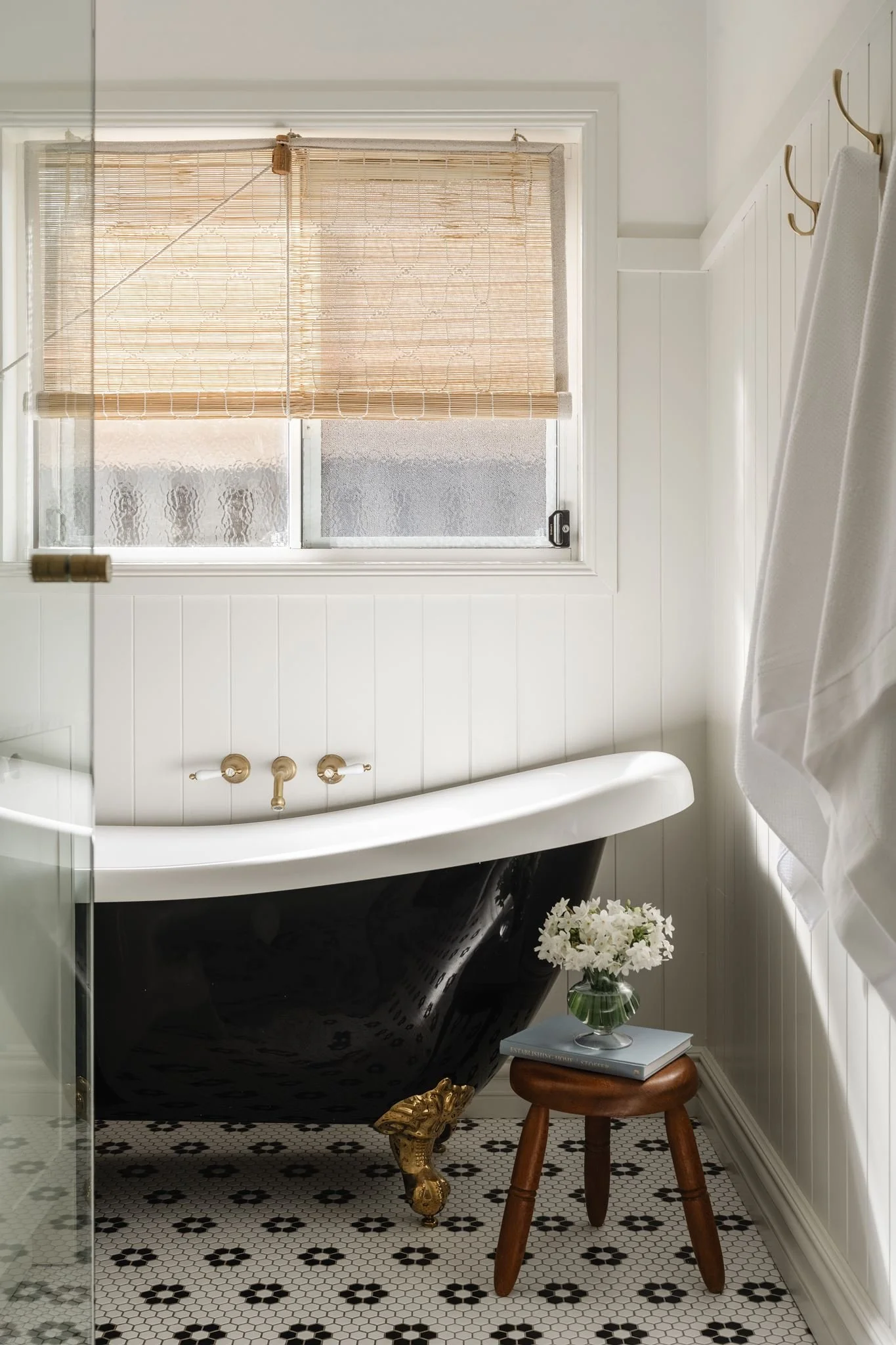 A bathroom with a white clawfoot bathtub with gold accents, a small wooden stool with a vase of white flowers, a white tiled wall, a window with a bamboo blind, and black and white hexagon tile flooring.