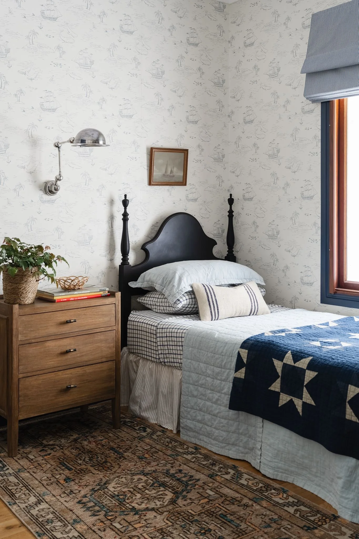 A bedroom with a black bed frame, white bedding, and checked pillows. Beside the bed is a wooden nightstand with a potted plant, books, and a small basket. A vintage painting hangs above the bed, and a wall-mounted, silver reading lamp is to the left