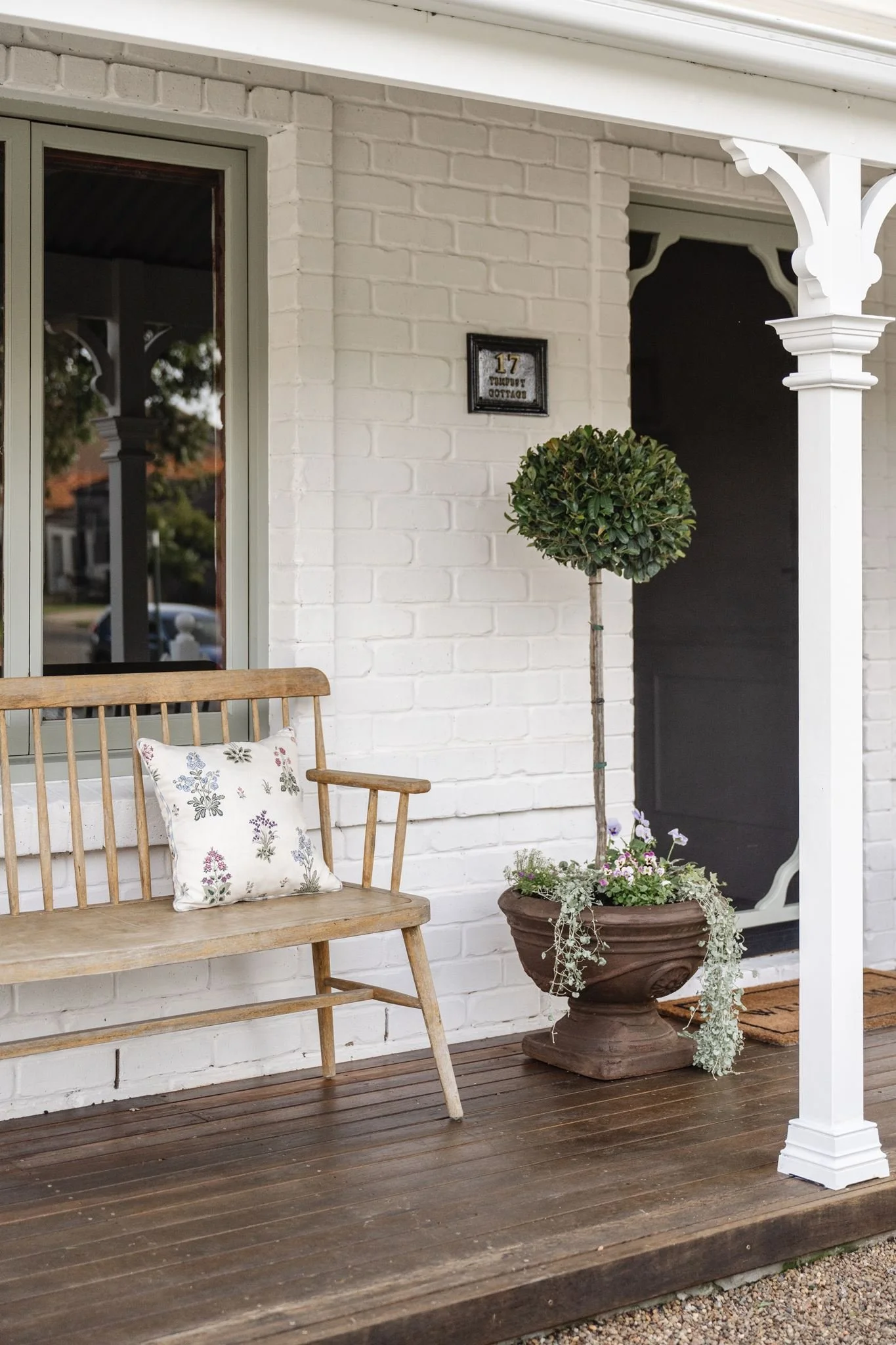 White brick house porch with a wooden bench, a floral pillow, a potted topiary tree, and a large decorative urn with plants. There is a window, a porch post, and a house number sign reading '17 Tempest Cottage'.