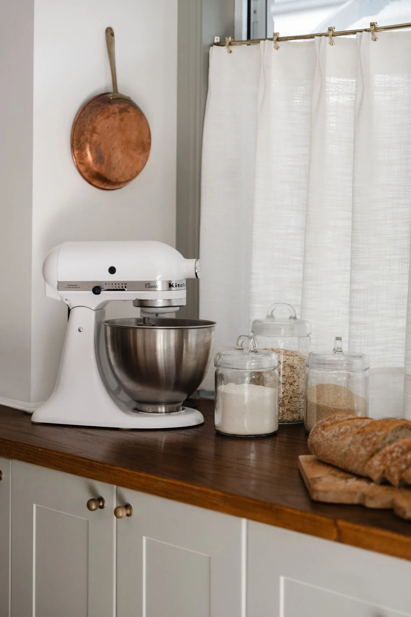 A kitchen countertop with a white stand mixer, four glass jars filled with flour, oats, and brown sugar, and a loaf of bread on a wooden cutting board. Part of a white cabinet with round knobs is visible at the bottom, and a white curtain hangs in fr