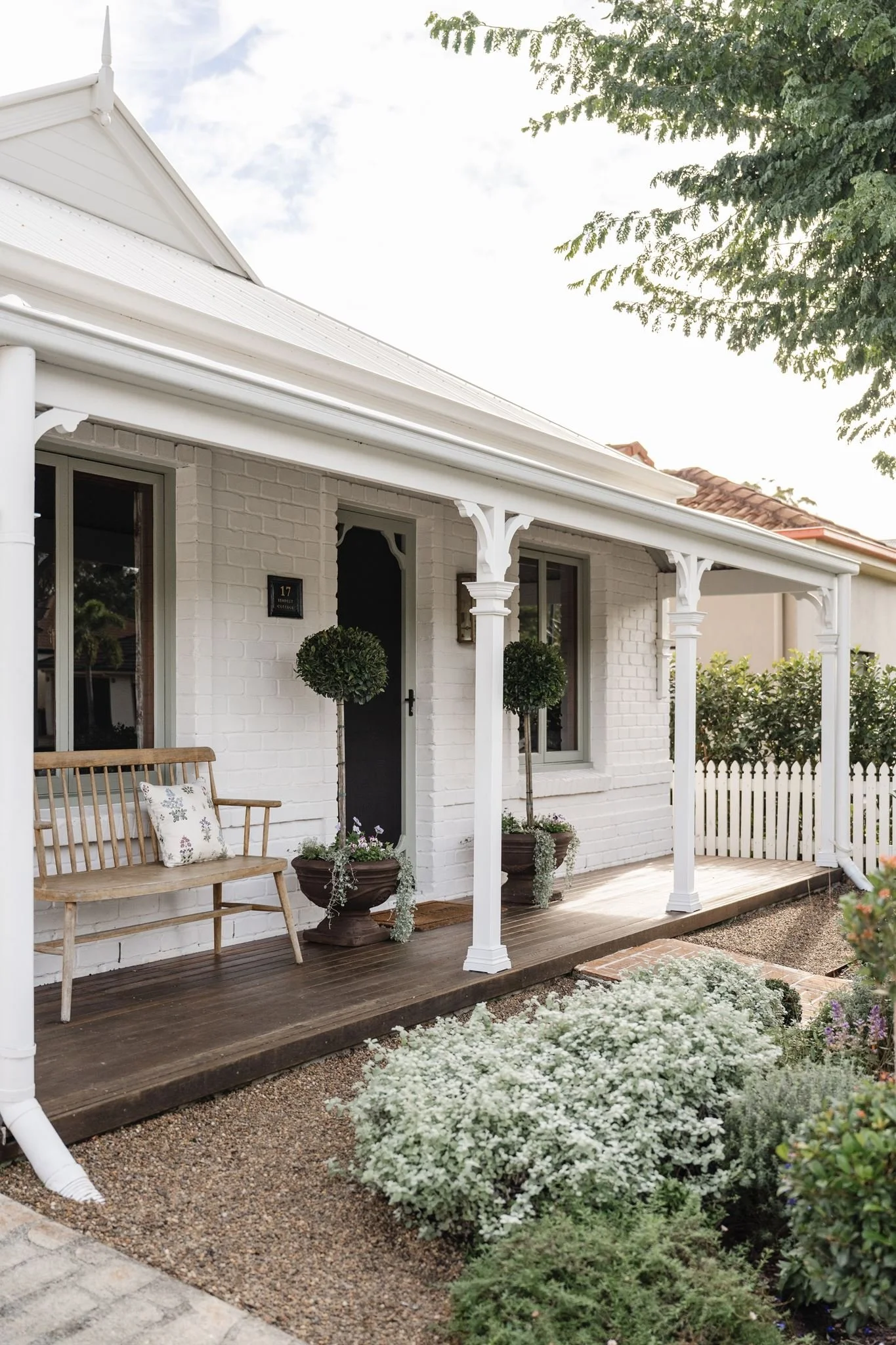 Front porch of a white brick house with a wooden deck, white columns, a wooden bench, potted plants, and decorative bushes in the garden.