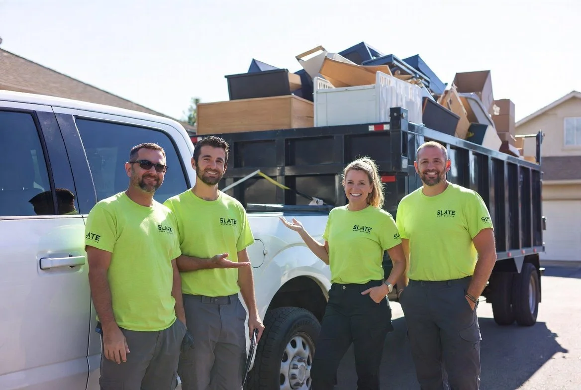 Four people stand in front of a moving truck filled with furniture, all wearing bright yellow-green shirts with a company logo, smiling on a sunny day.
