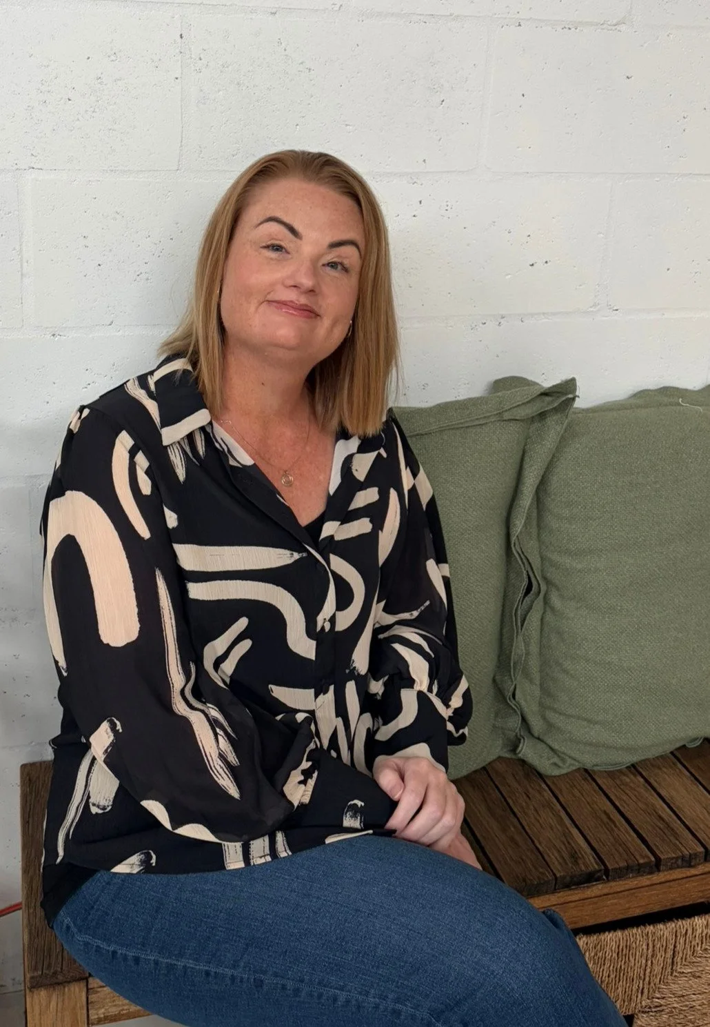 A woman with shoulder-length light brown hair and fair skin, wearing a black shirt with beige abstract patterns, sitting on a wooden bench with green cushions against a white brick wall.