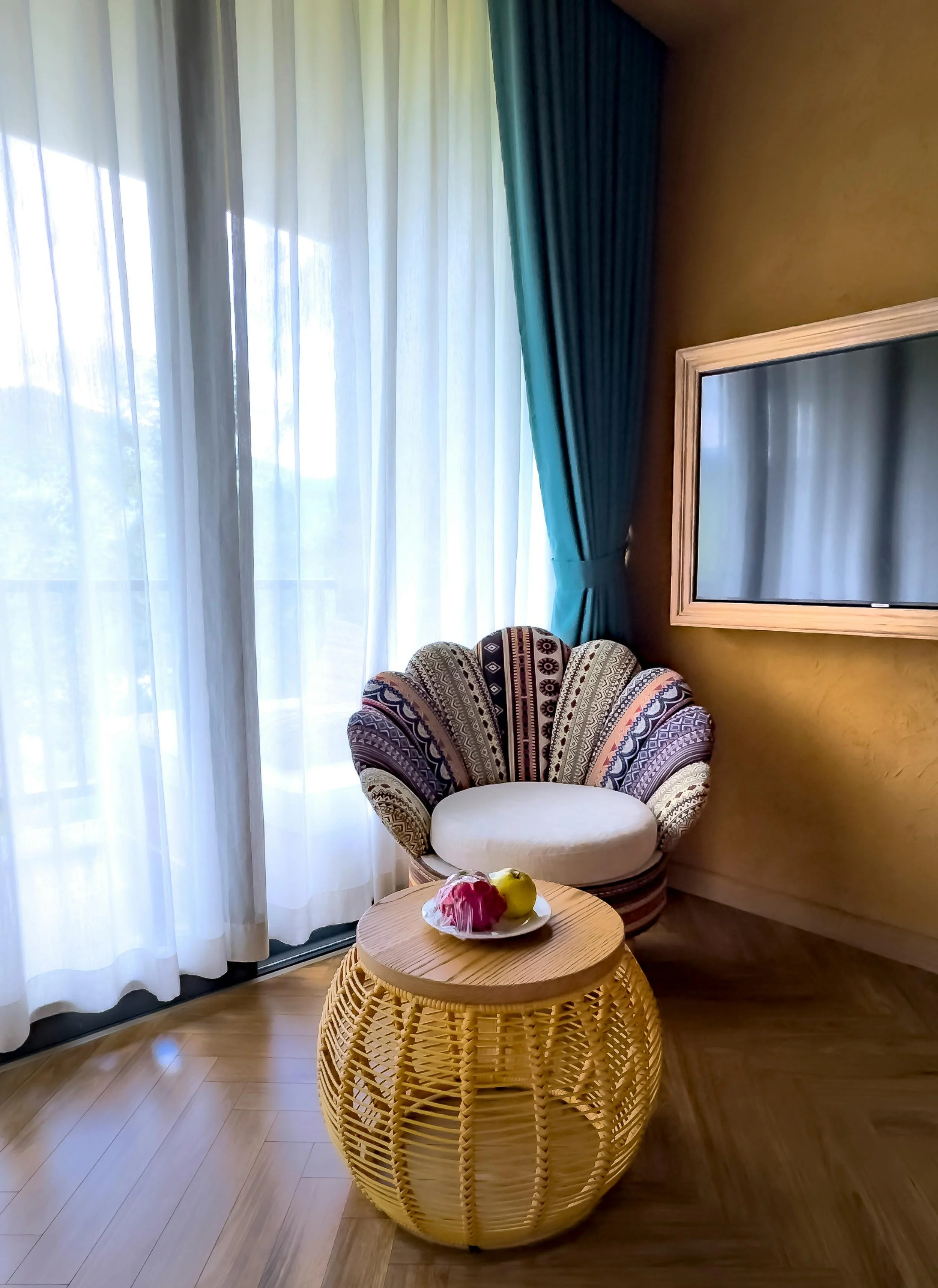 A cozy corner with sheer white curtains and blue drapes covering a glass door or window. A patterned armchair with a rounded back and cushion is next to the wall. A round wooden coffee table with a decorative wicker base holds a bowl of assorted fruits, including a red bell pepper and yellow apple. A large flat-screen TV is mounted on the wall with a light-colored wooden frame. The room has light wood flooring.