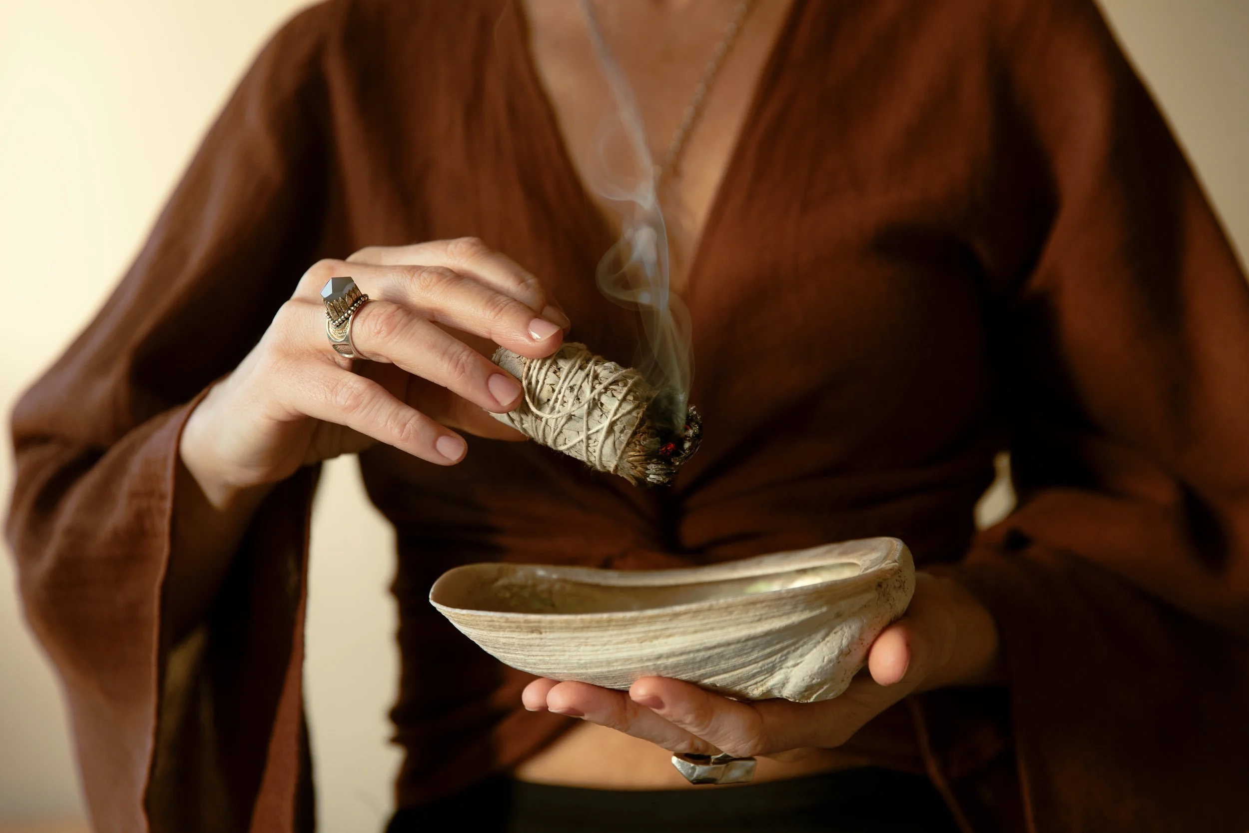 Person with brown top holds a burning sage stick over a beige ceramic bowl.
