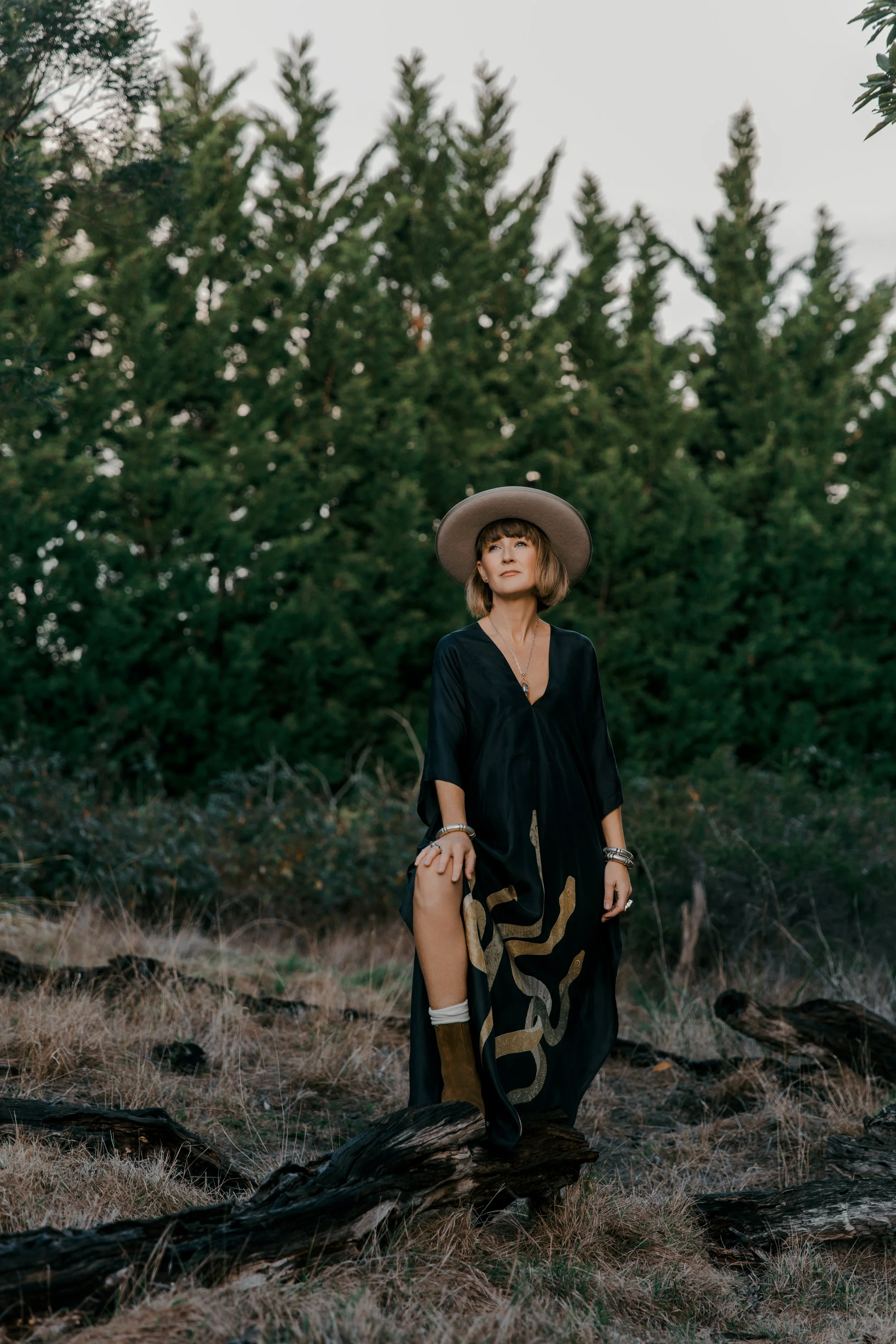 A woman wearing a wide-brimmed hat and a black dress standing outdoors on a log amidst a natural setting with tall trees and dry grass.
