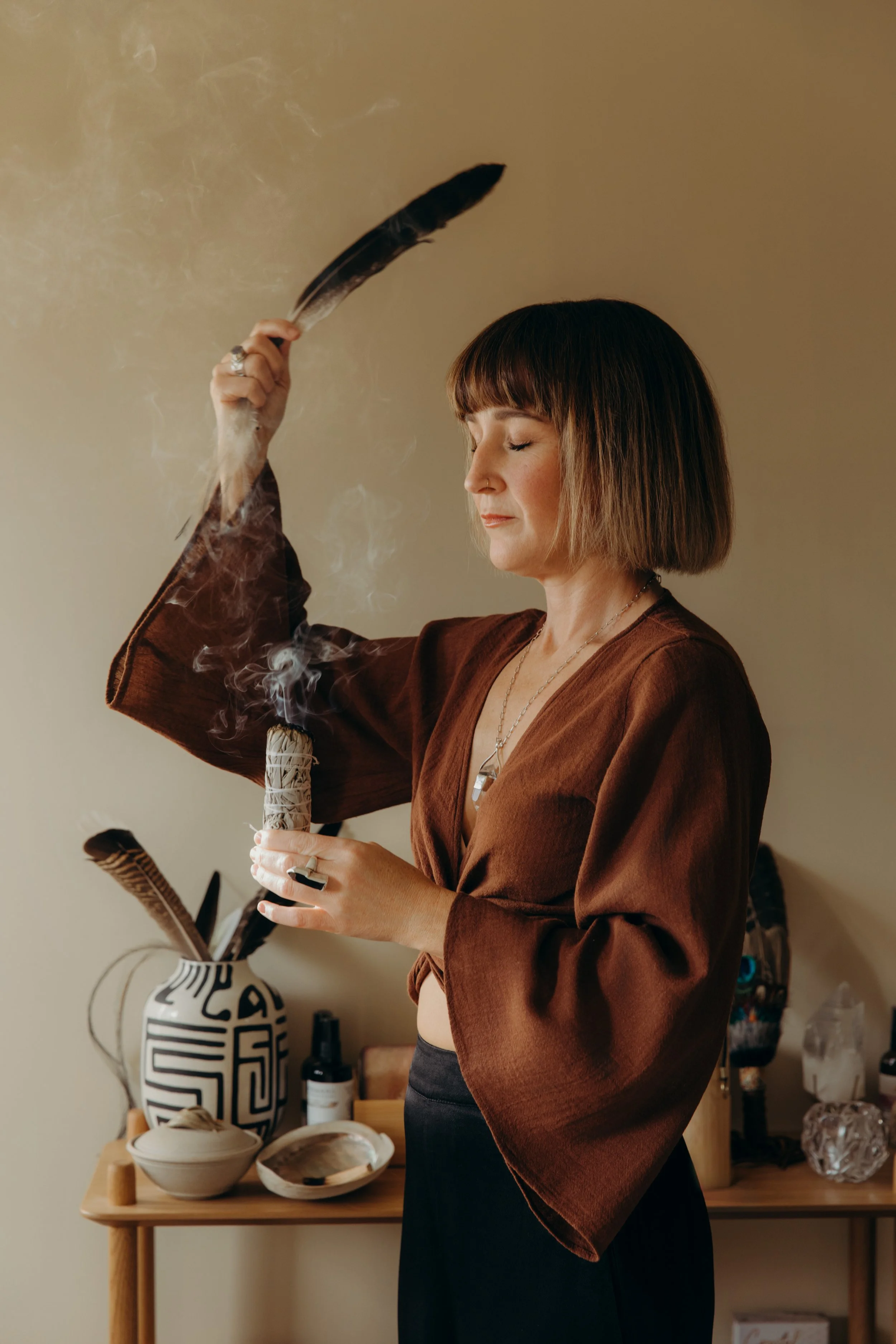 A woman with shoulder-length brown hair and a nose piercing is holding a burning smudge stick and appears to be in a meditative or relaxing state with her eyes closed. She is standing in front of a wooden table with various bowls and vases, some containing feathers, and plants in a decorative vase.