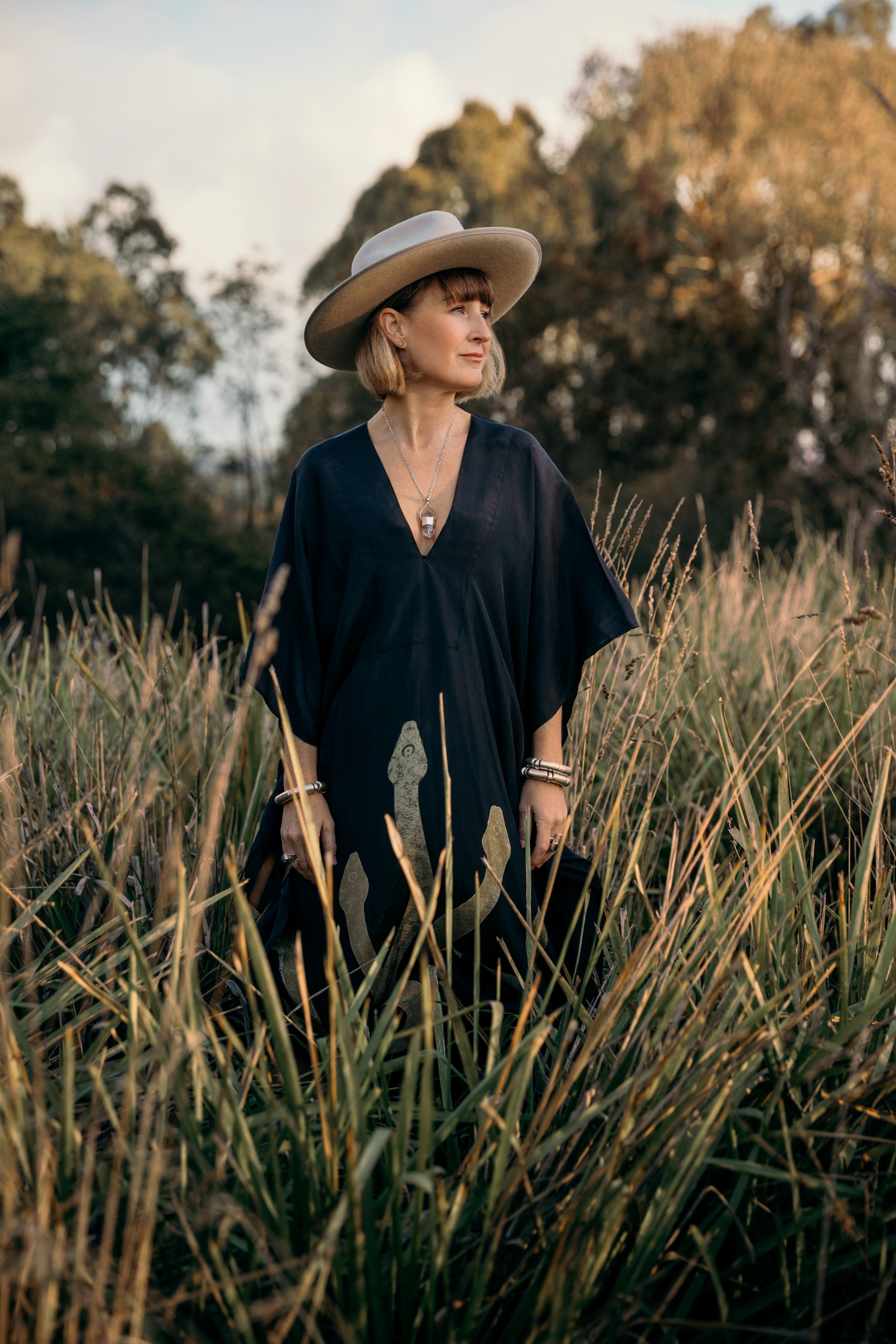 Woman in a wide-brimmed hat and dark dress standing in a field of tall grass with trees in the background during golden hour.