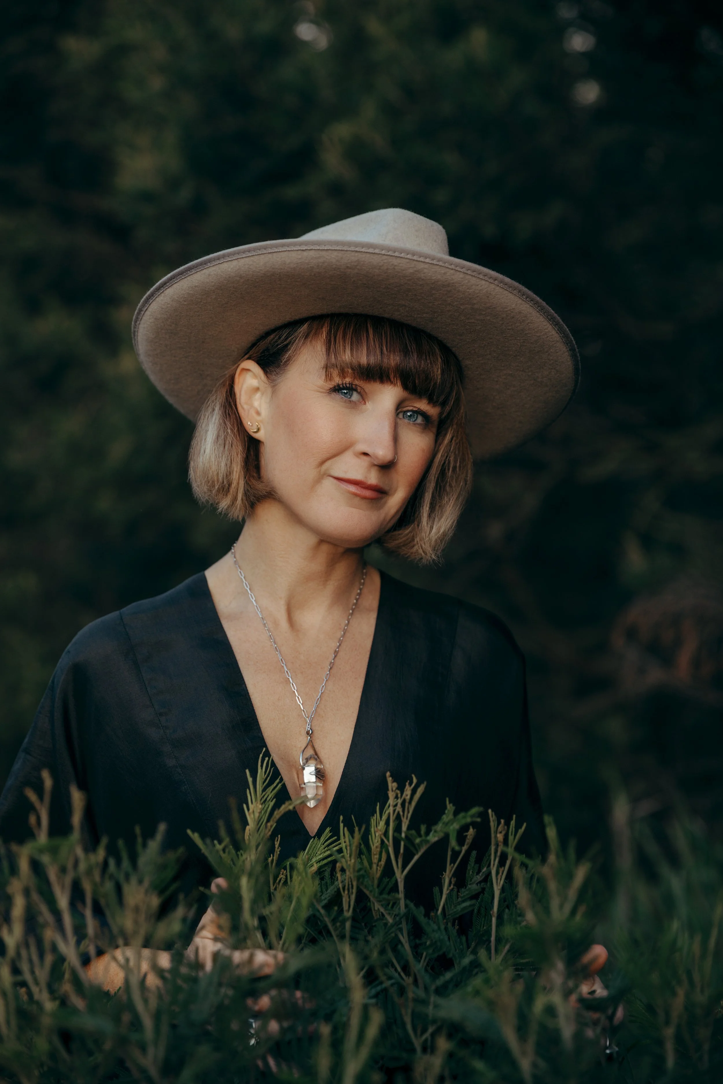 A woman with short brown hair, blue eyes, and multiple earrings, wearing a large beige hat and a black blouse with gold embroidery, stands in a forested area with green foliage.