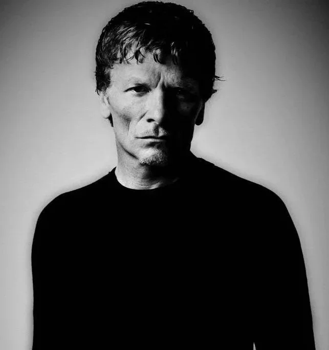 Black and white portrait of a man with short, wavy hair, wearing a dark shirt, looking serious and staring directly at the camera.