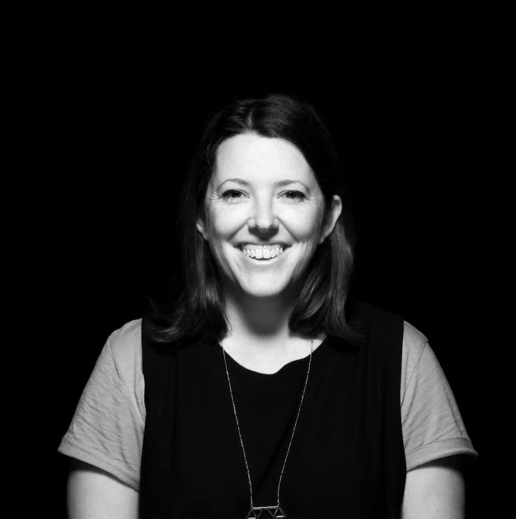 Black and white portrait of a smiling woman with shoulder-length hair, wearing a dark top and a necklace, against a black background.