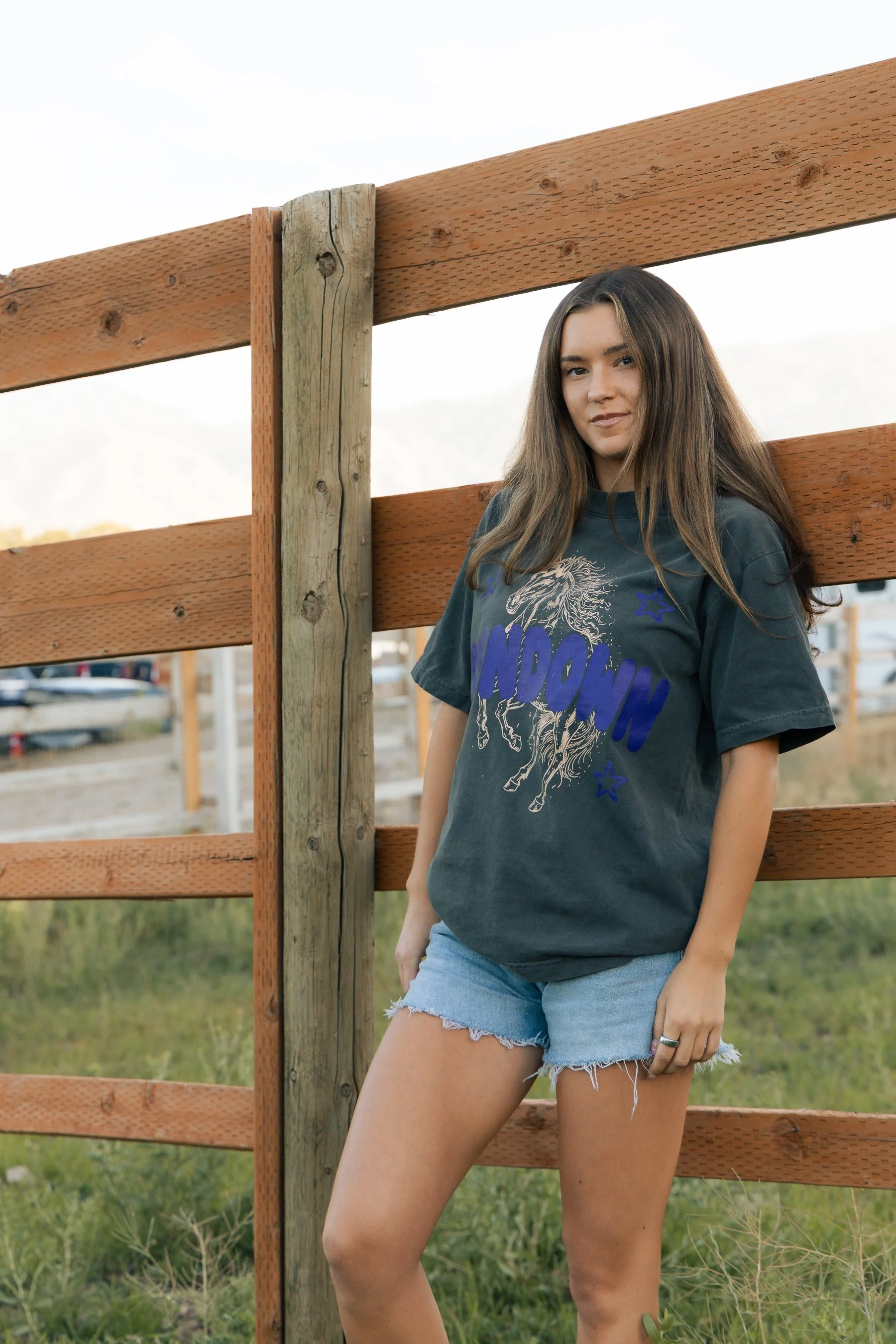 A young woman with long brown hair standing outdoors next to a wooden fence, wearing a dark t-shirt with a graphic design and light denim shorts, in a grassy area.
