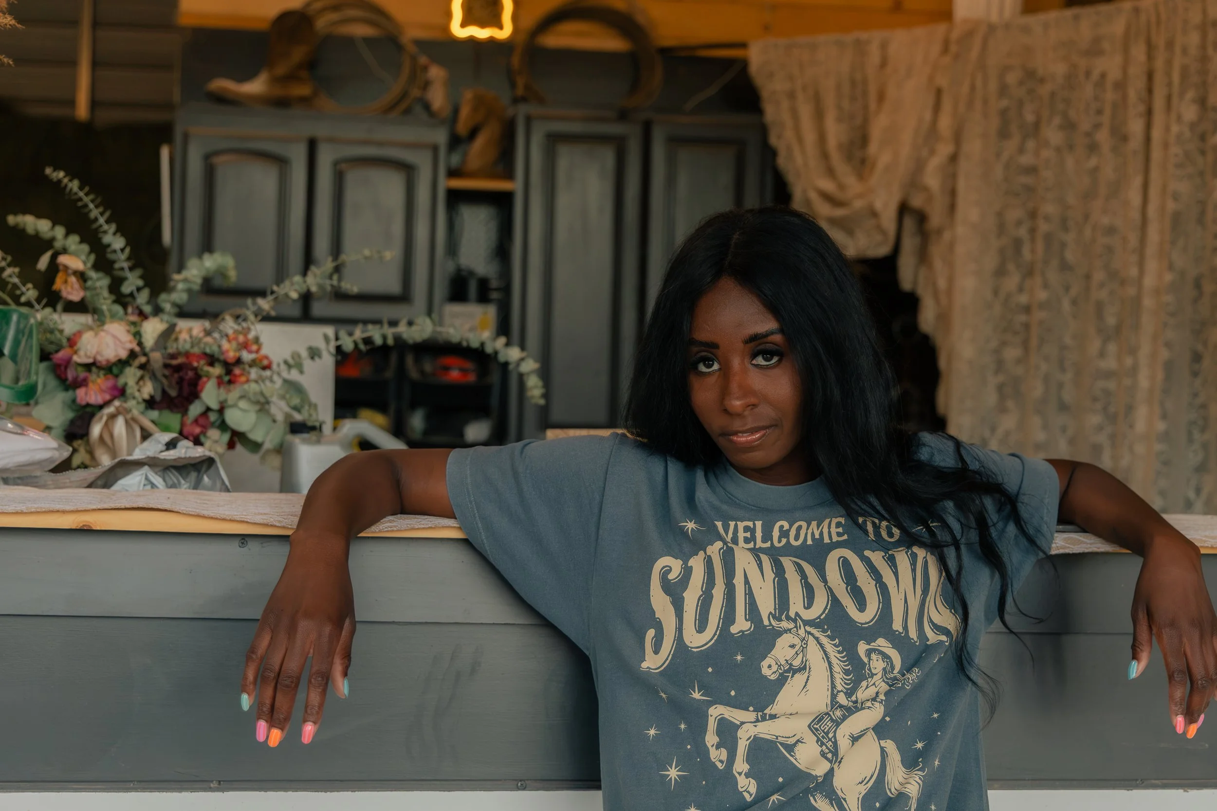 A woman with long dark hair and dark eyes leaning on a table with holding her arms out, showing her colorful manicured nails. She is wearing a blue T-shirt with a graphic design. In the background, there is a shelf with decorations, flowers, and hanging supplies in a cozy, rustic setting.
