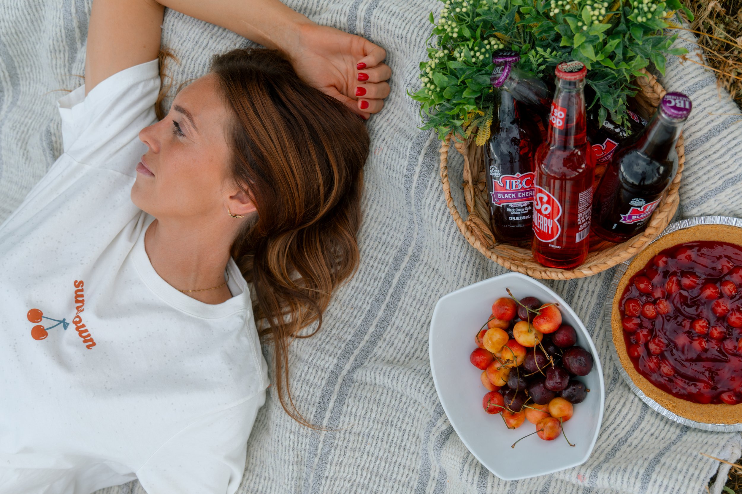 A woman with reddish-brown hair and red nails lying on a blanket outdoors surrounded by bowls of cherries and a pie with cherry topping, with a basket of cherry soda bottles nearby.