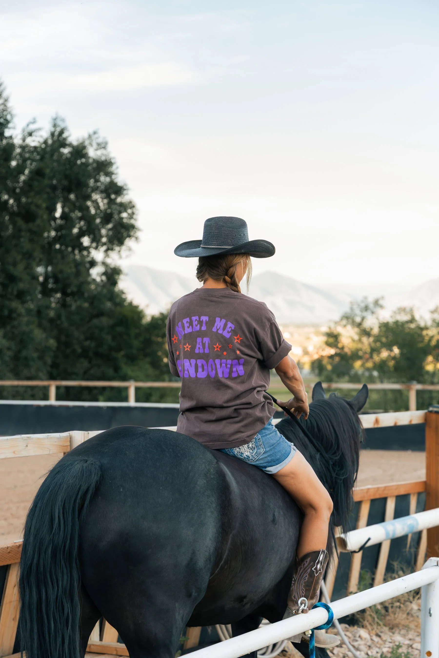A woman wearing a wide-brimmed hat and a t-shirt that reads 'Meet Me at Sunset' rides a black horse inside a fenced outdoor riding arena with trees and mountains in the background.