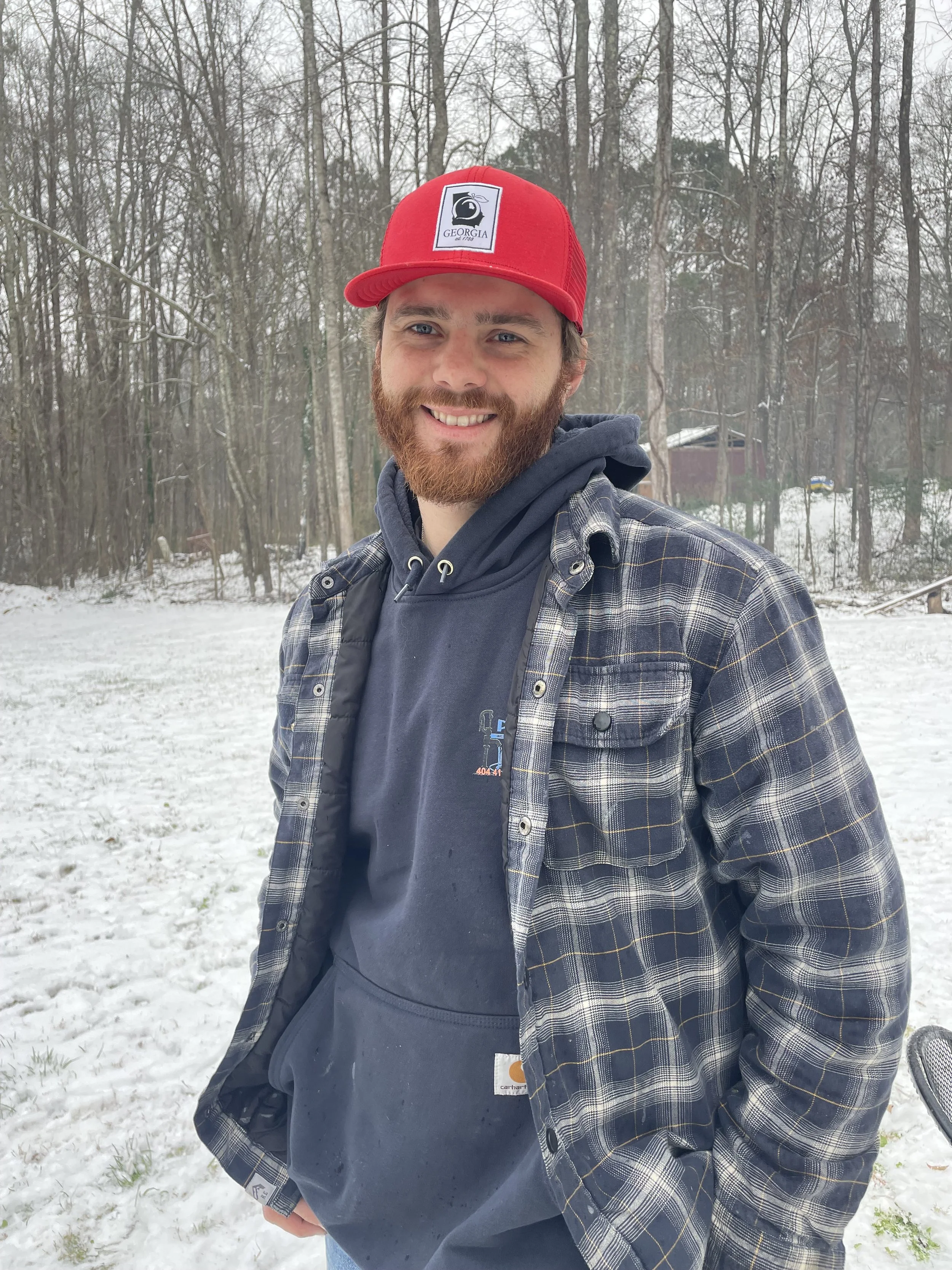 A man with a beard standing outside in a snowy landscape, wearing a red Georgia baseball cap and a layered outfit including a checkered jacket and a hoodie.