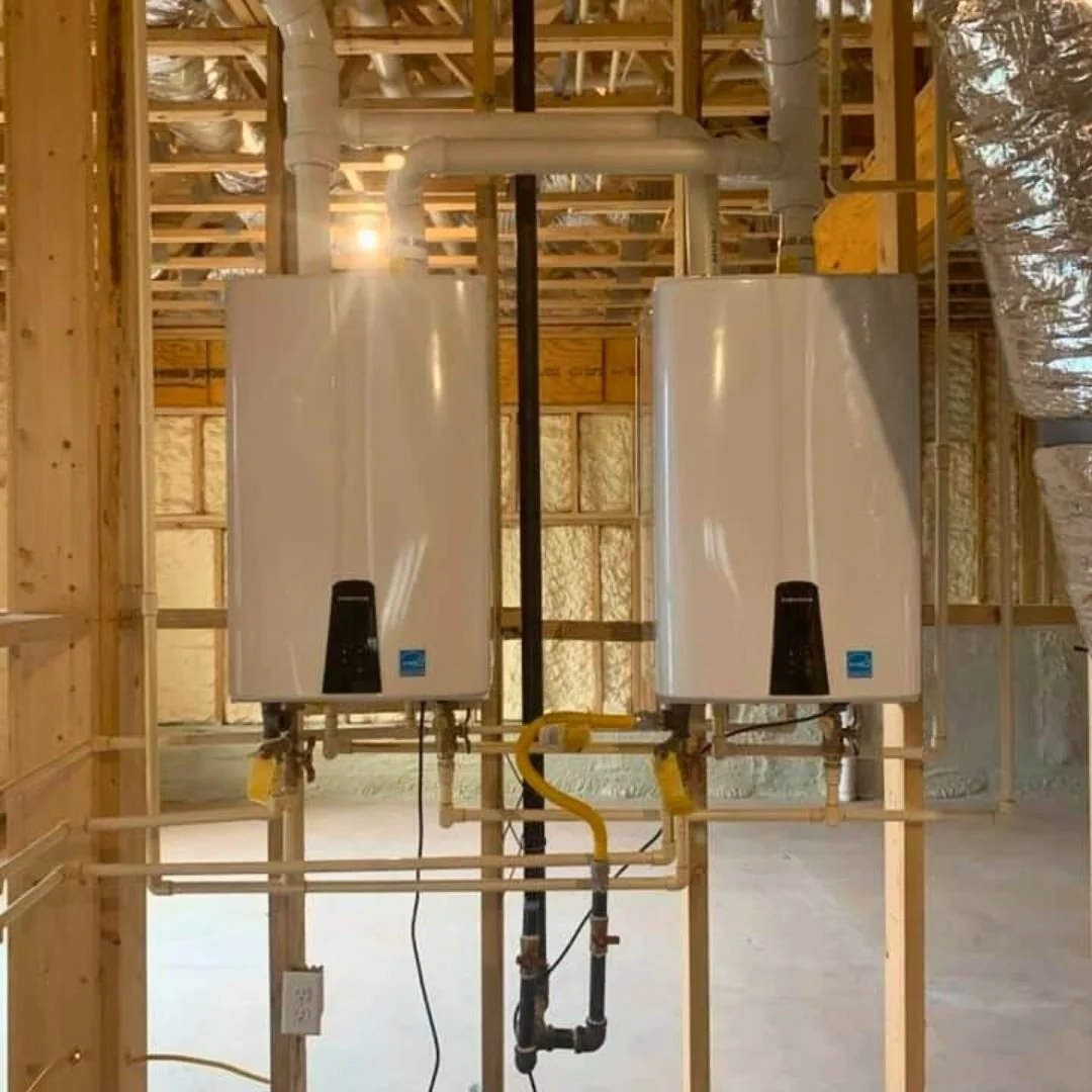 Two white tankless water heaters mounted on a wooden wall in a building under construction, with pipes and venting systems visible.