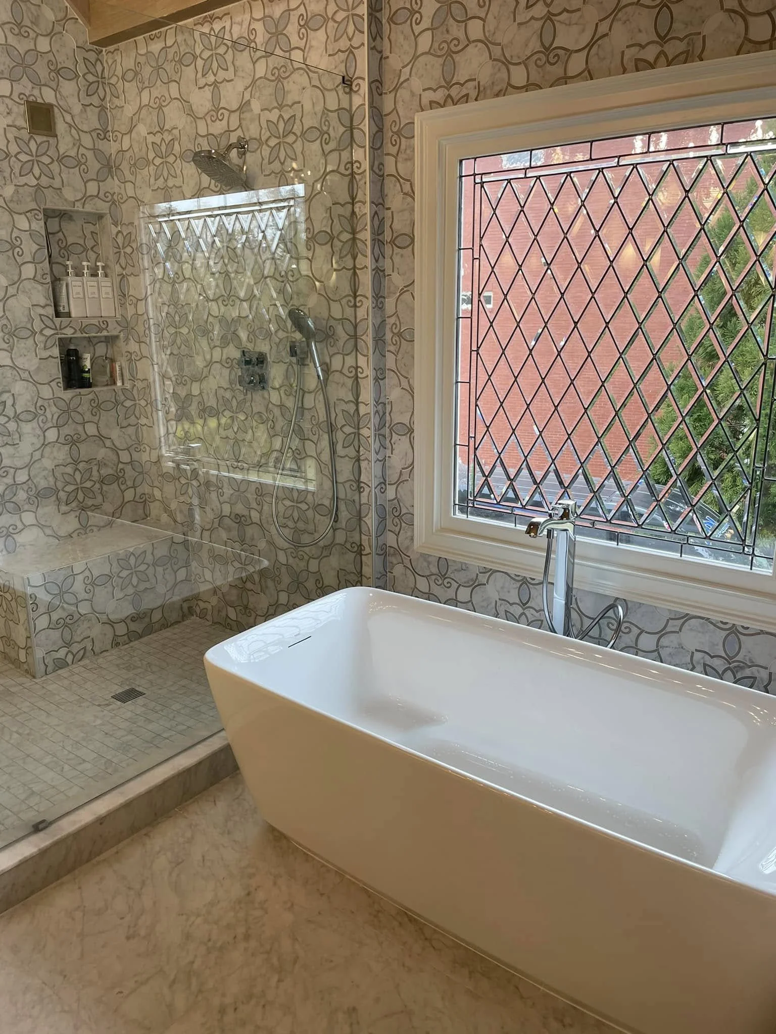 A bathroom with a large window secured by black metal grilles, a freestanding bathtub, and a walk-in shower area with a patterned tiled wall and built-in shelves for toiletries.