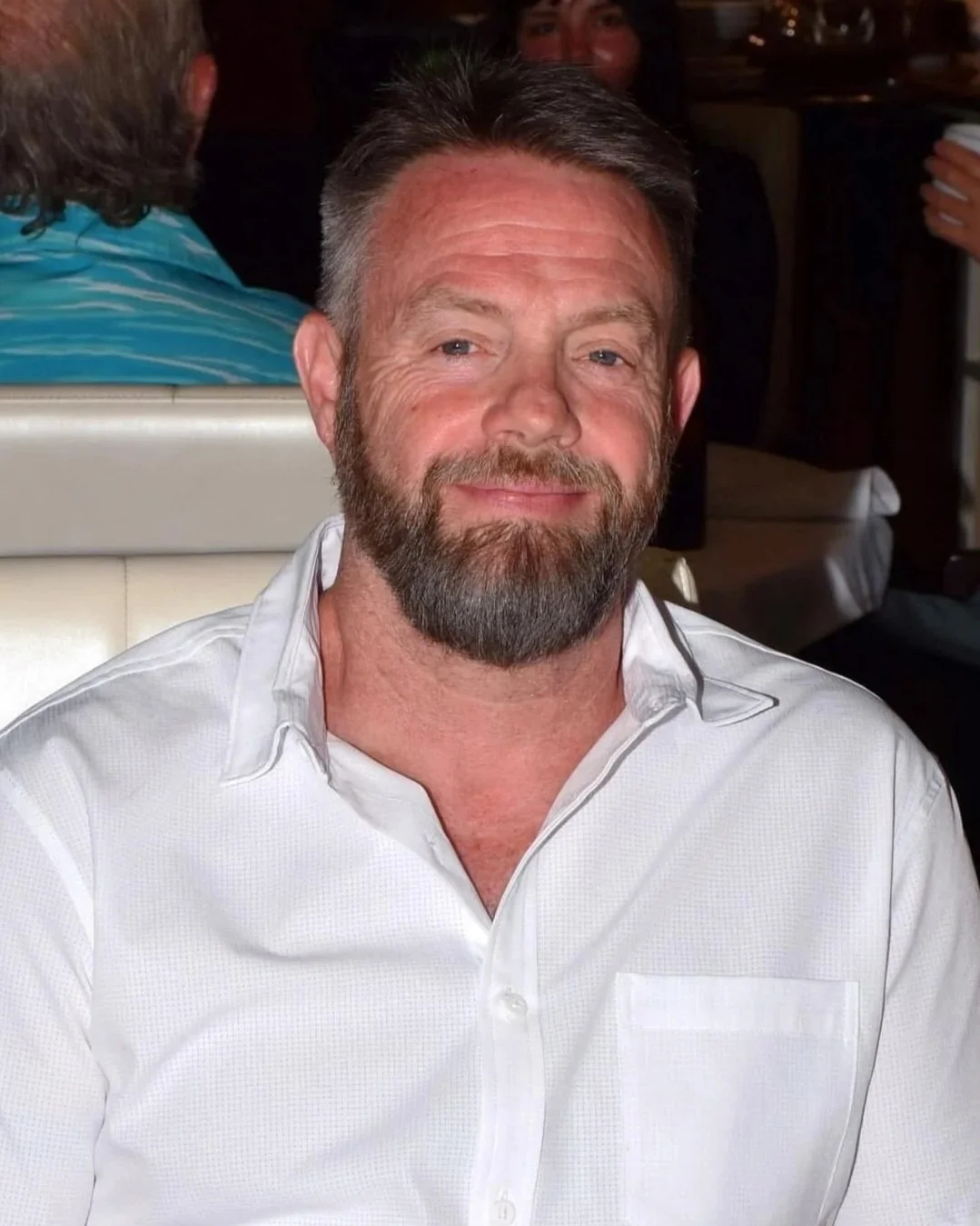 A man with gray hair and beard, wearing a white dress shirt, smiling at a social gathering.