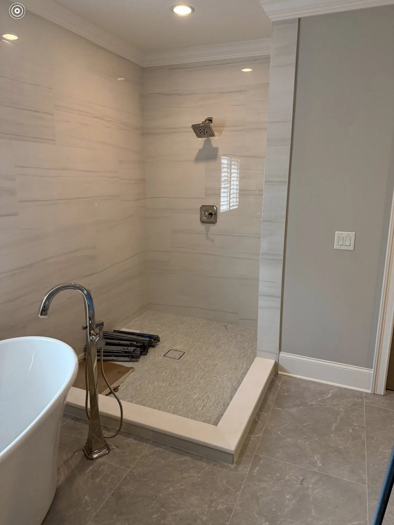 A bathroom with a large walk-in shower with beige tiled walls, a rain showerhead, a wall-mounted control, and a drain on the floor. There is a white bathtub in the foreground, with a silver faucet and handheld shower attachment. Several black foldabl