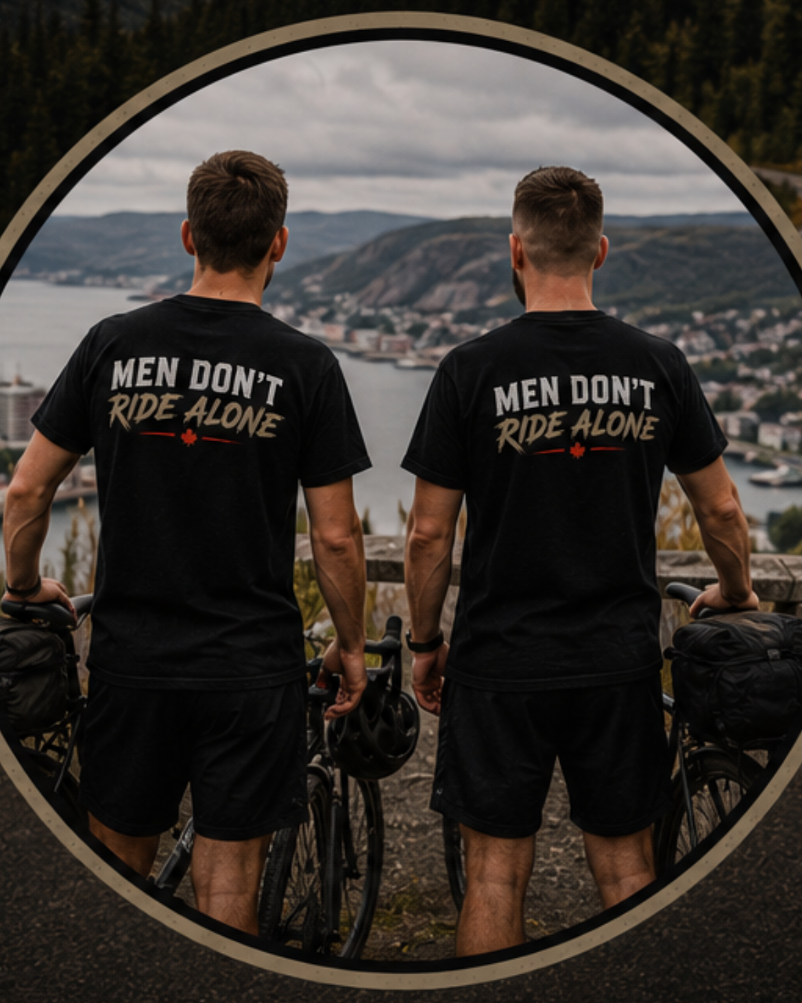 Two men standing with bicycles wearing matching black T-shirts that say "Men Don't Ride Alone" on the back. They are overlooking a scenic view of a river, hills, and a town under a cloudy sky.