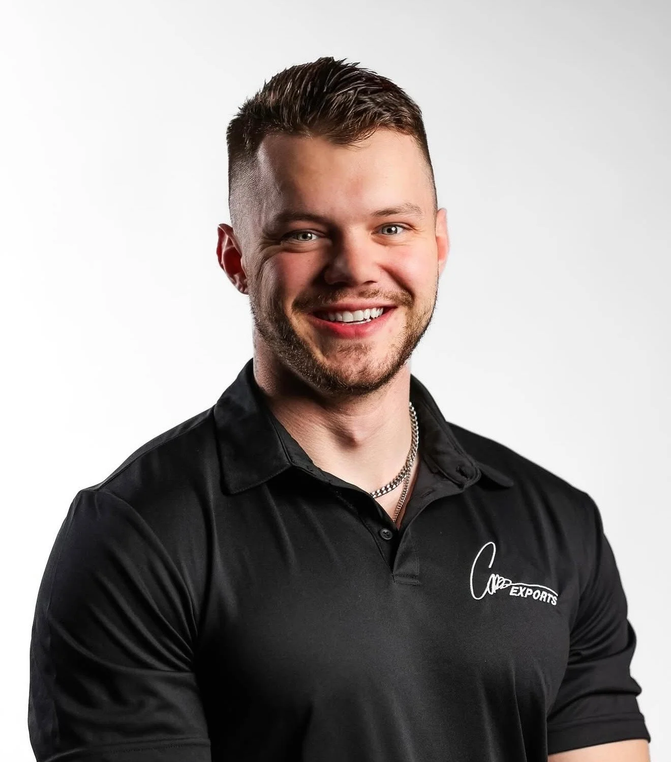 A young man with short brown hair, beard, and blue eyes, smiling, wearing a black polo shirt with a logo and silver chain necklace, standing against a plain white background.