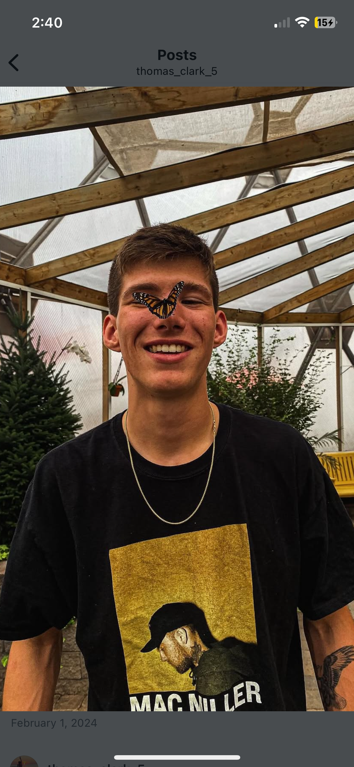 A young man with short brown hair and a big smile in a greenhouse or garden structure, with a butterfly perched on his nose. He is wearing a black t-shirt with a graphic print and a silver chain necklace.