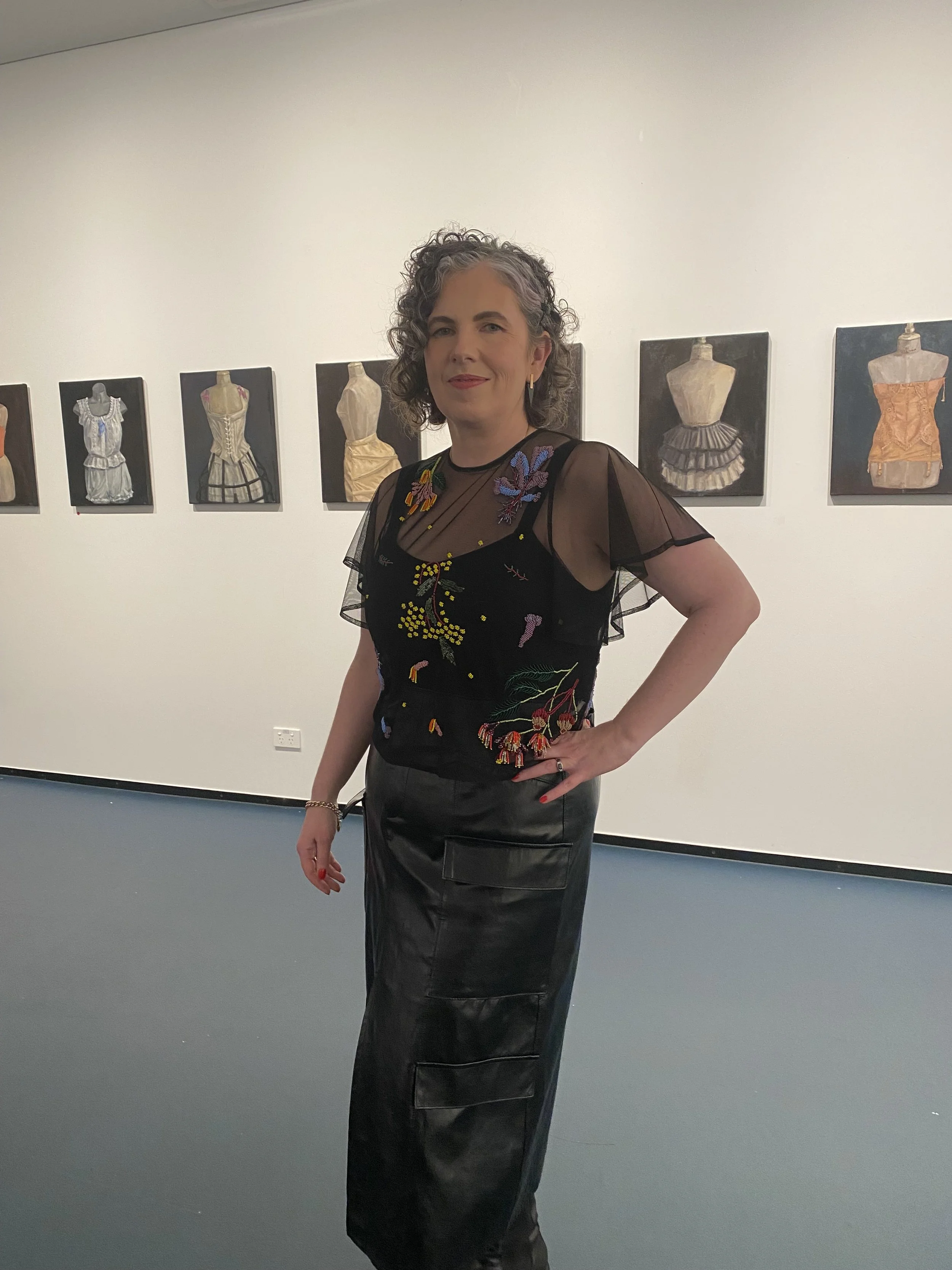 photo of a woman dressed in black standing in a gallery in front of a series of paintings of mannequins in vintage corsetry and undergarments