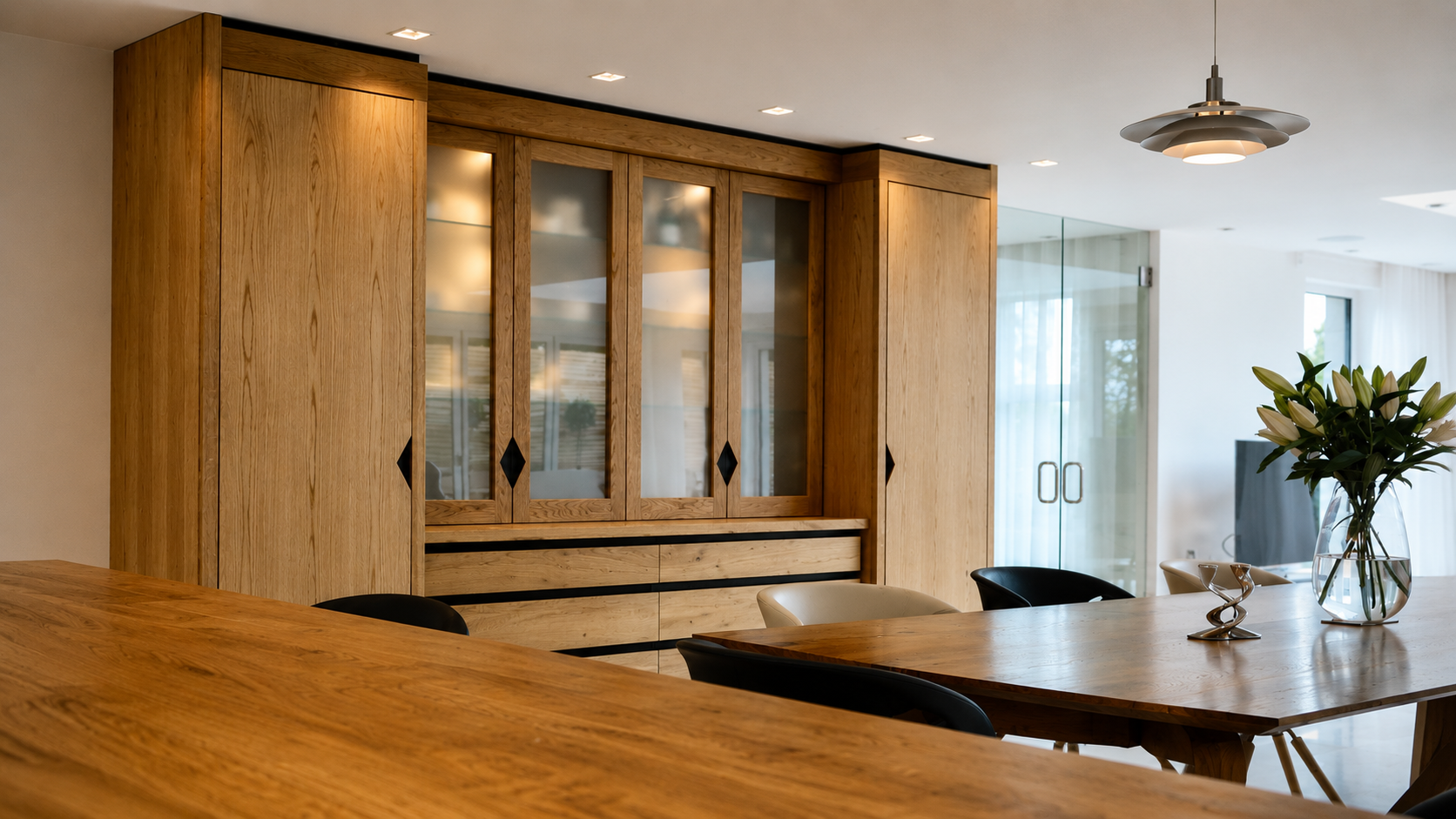 Modern kitchen with wooden cabinets, glass-paneled doors, a dining table, and a vase of white lilies.