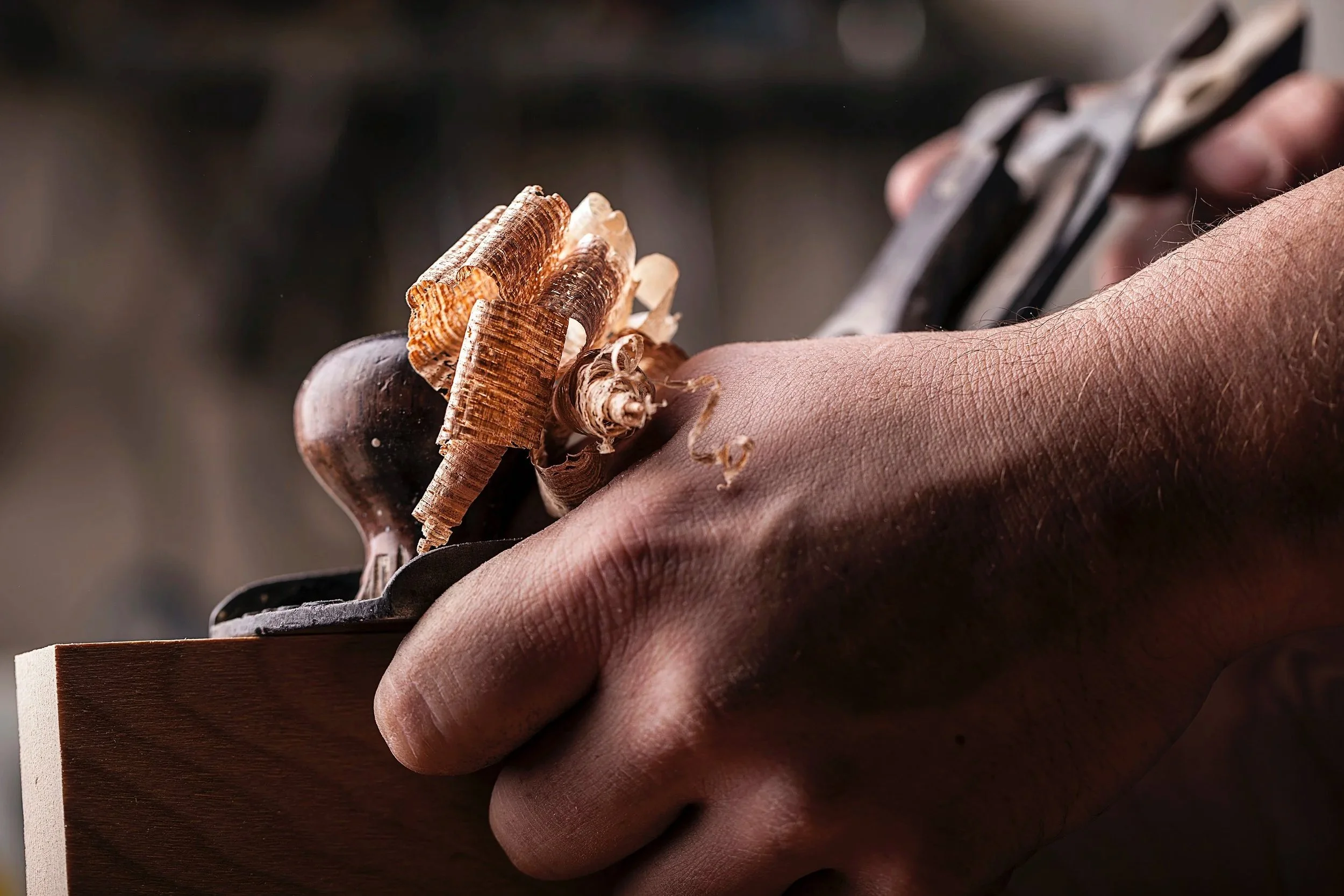 Close-up of a Nathan's hand holding a woodworking plane, smoothing a piece of wood, with wood shavings on top.