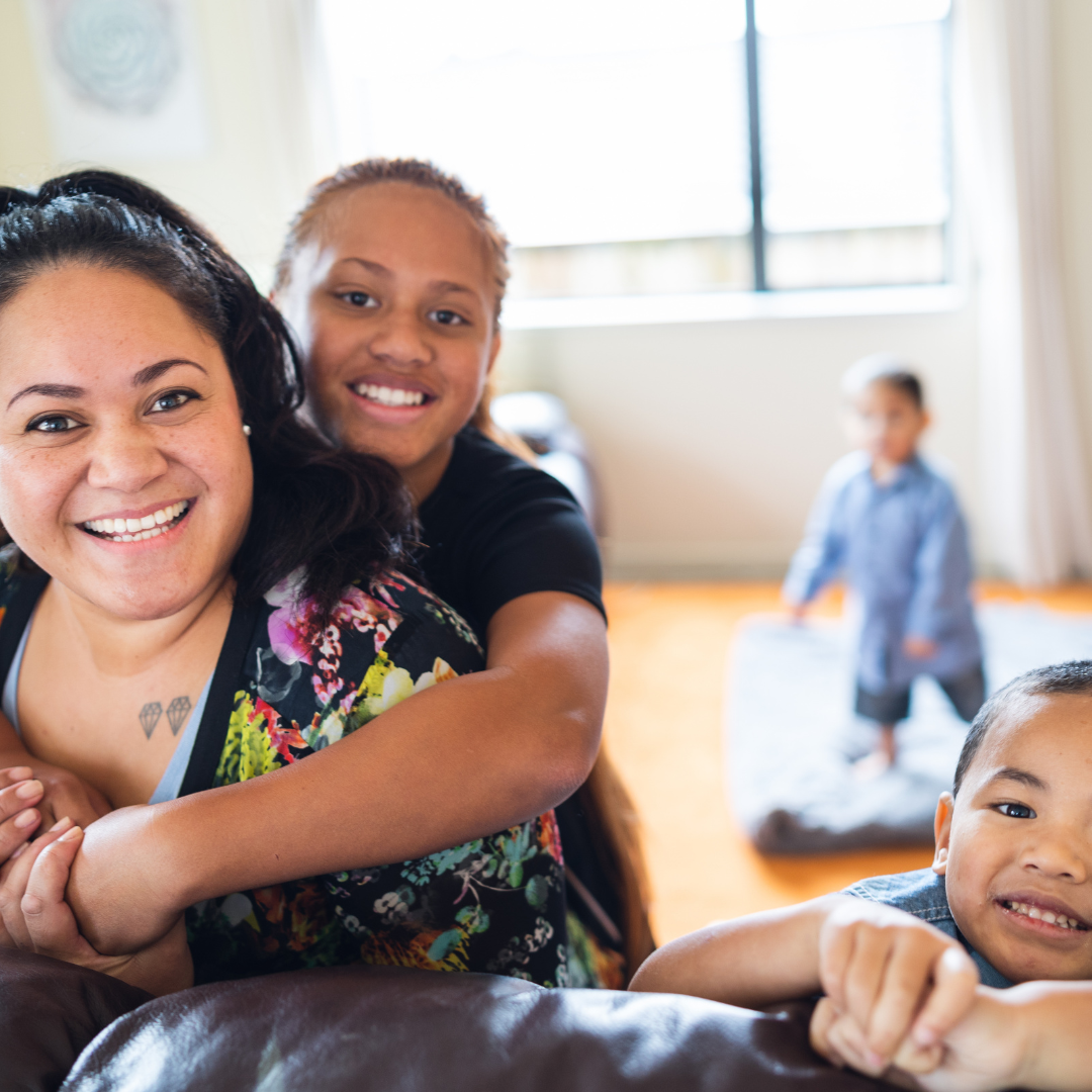 A woman and two children smiling and playing in a bright living room, with a third child in the background.