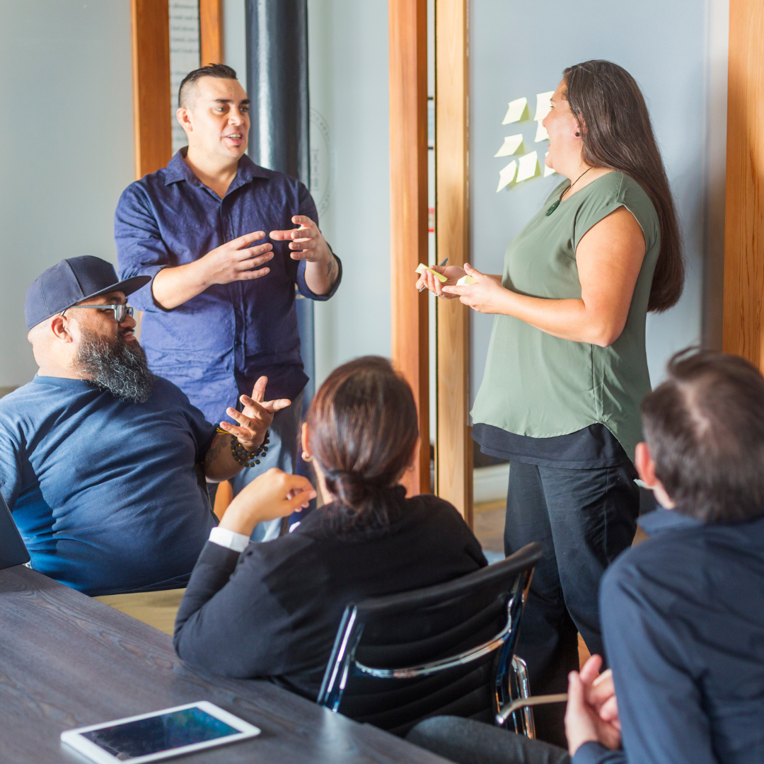 Group of diverse people in a business meeting, with one woman standing and speaking to the seated group, some taking notes and listening attentively.