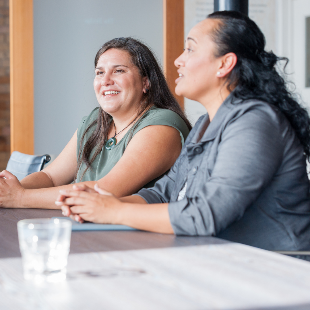 Two women sitting at a table in a meeting room, smiling and engaging in conversation.