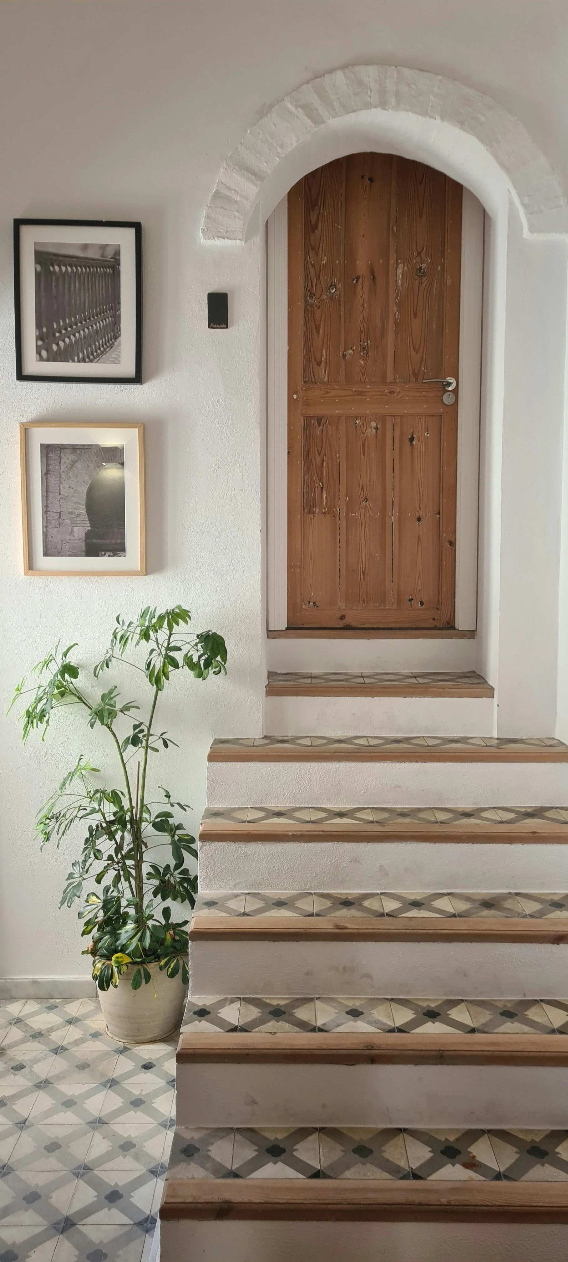 Interior entryway with a staircase, wooden door, potted plant, and framed black-and-white photographs on white wall.