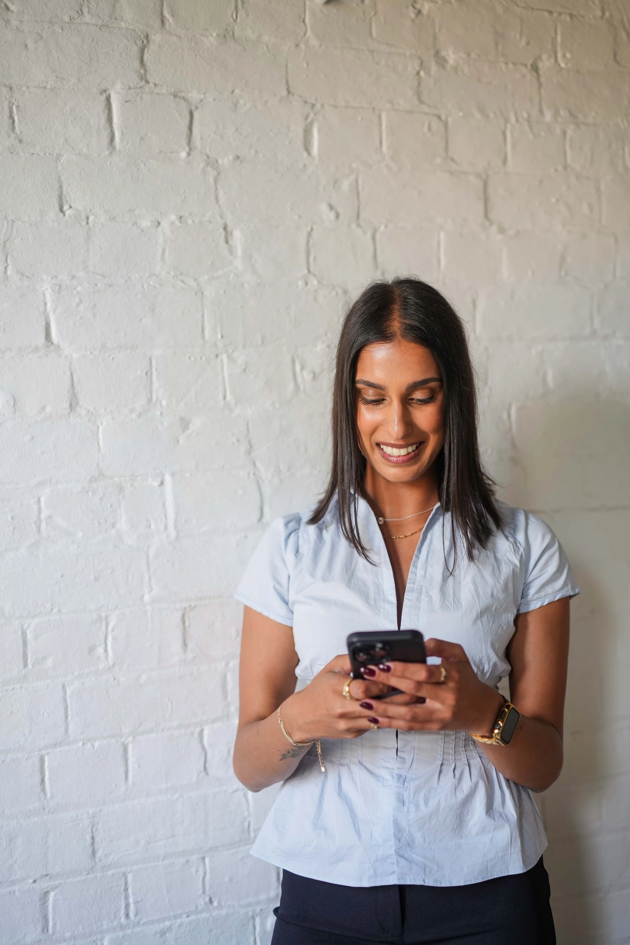 A woman with long dark hair, wearing a light blue blouse and black pants, standing against a white brick wall, smiling and looking at her smartphone.