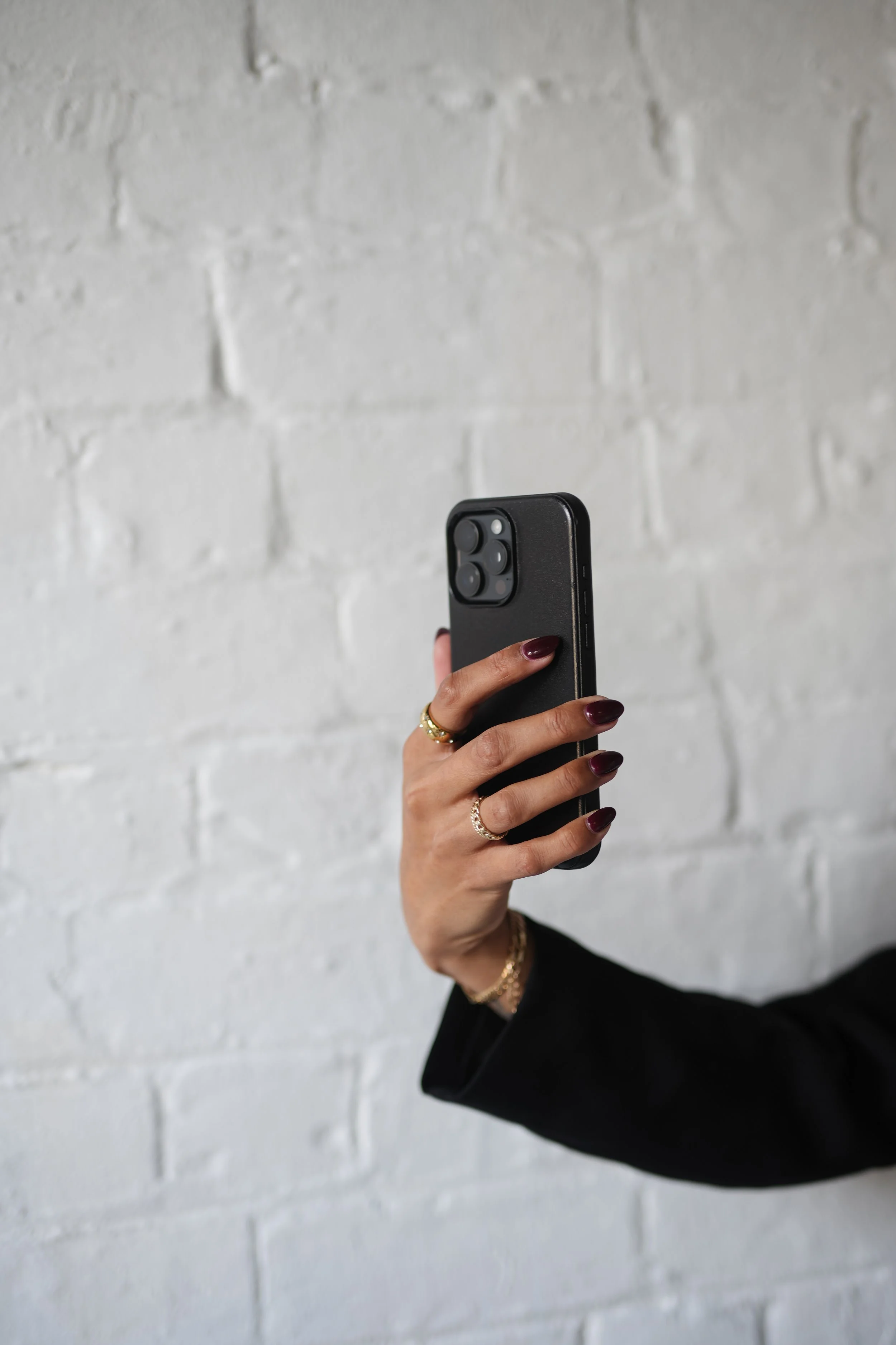 Close-up of a person holding a black smartphone with a triple-lens camera, against a white brick wall. The person's hand has manicured nails and they are wearing gold rings and bracelets, with a black sleeve visible.