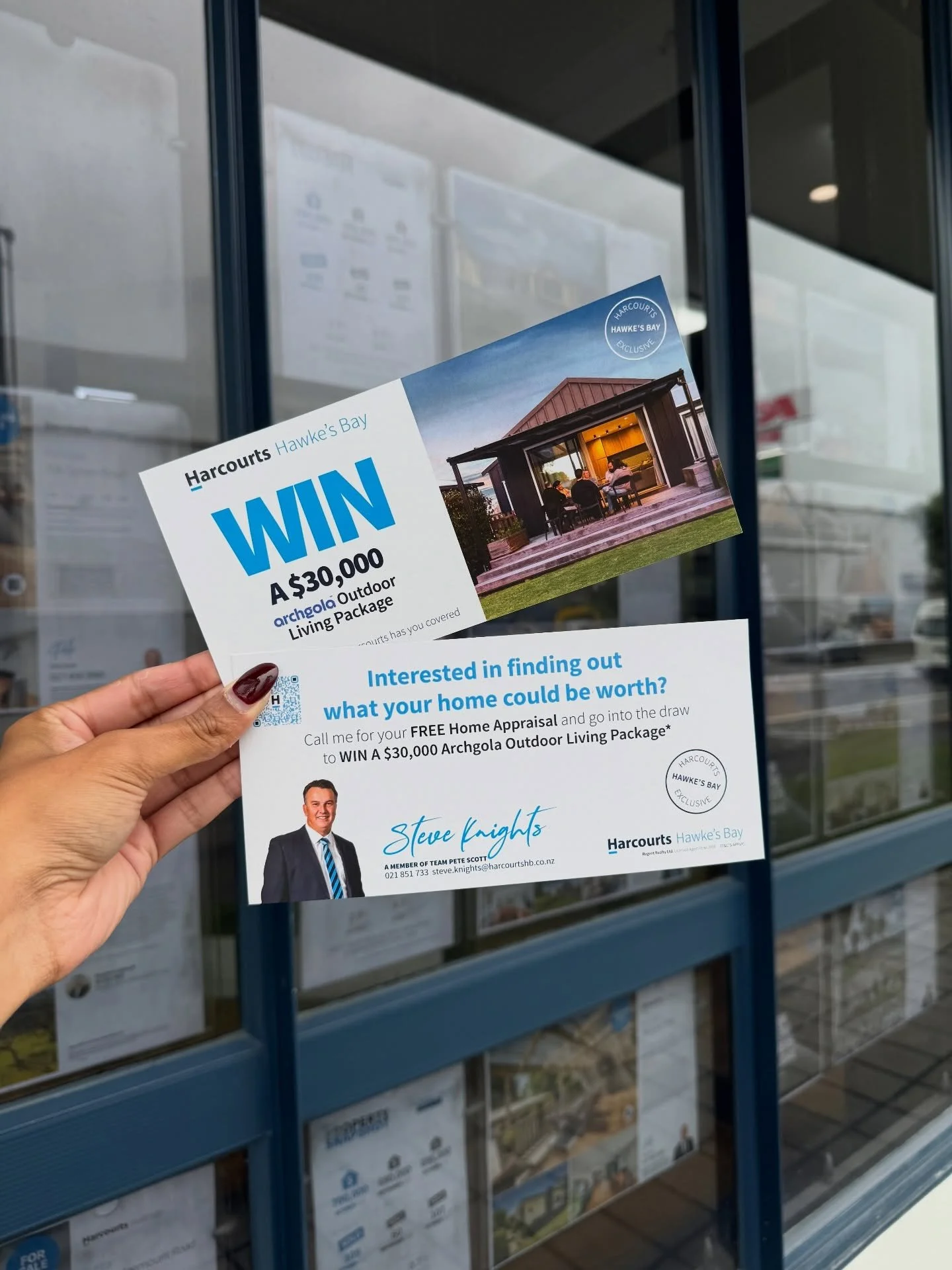 A Harcourts flyer held in front of a building window advertises a contest to win a $30,000 outdoor living package, featuring a house with an outdoor seating area and a photo of a man named Steve Knights.