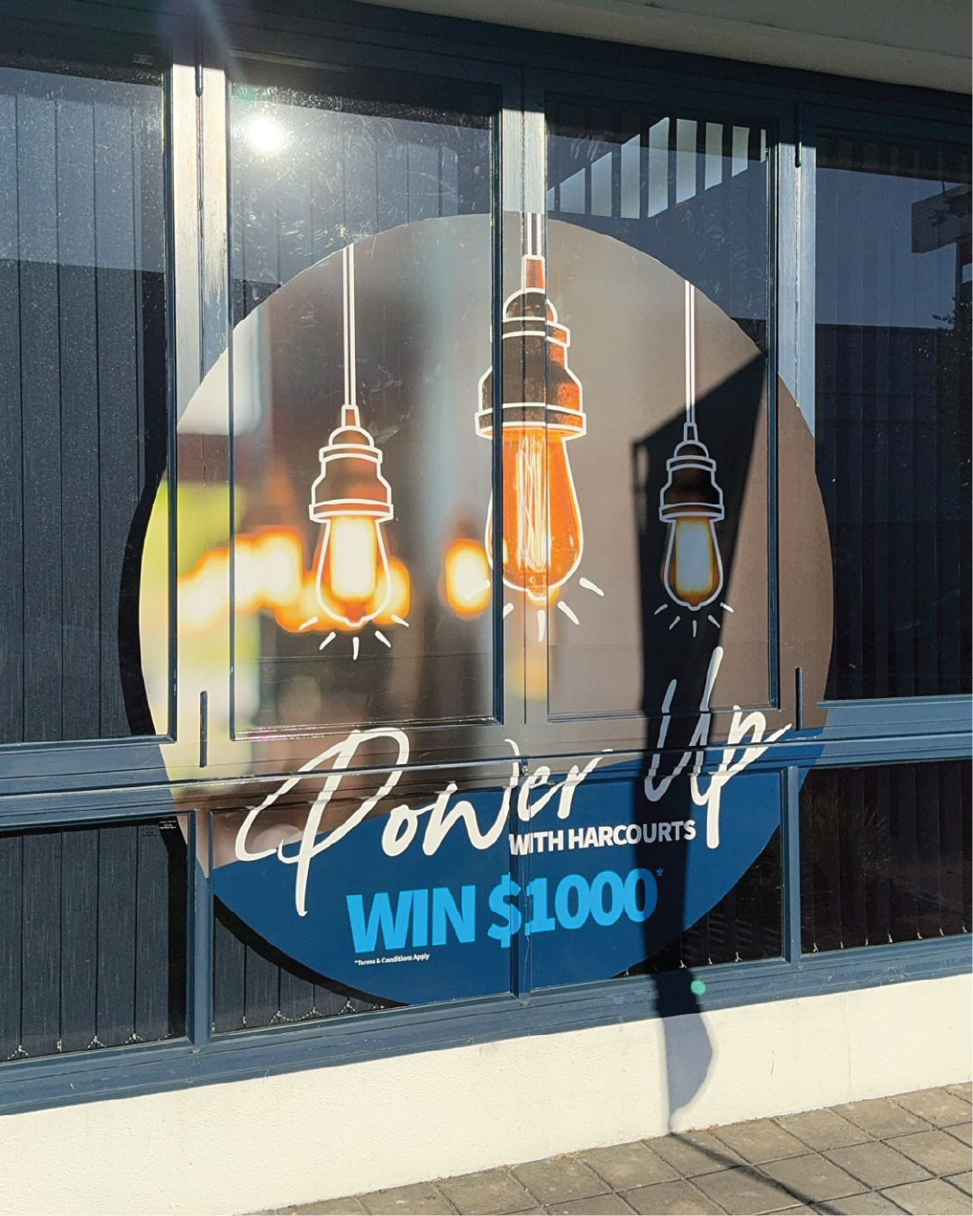 Marketing campaign collateral. The sign promotes a contest offering a chance to win $1,000, with the wording 'Power Up with Harcourts'.