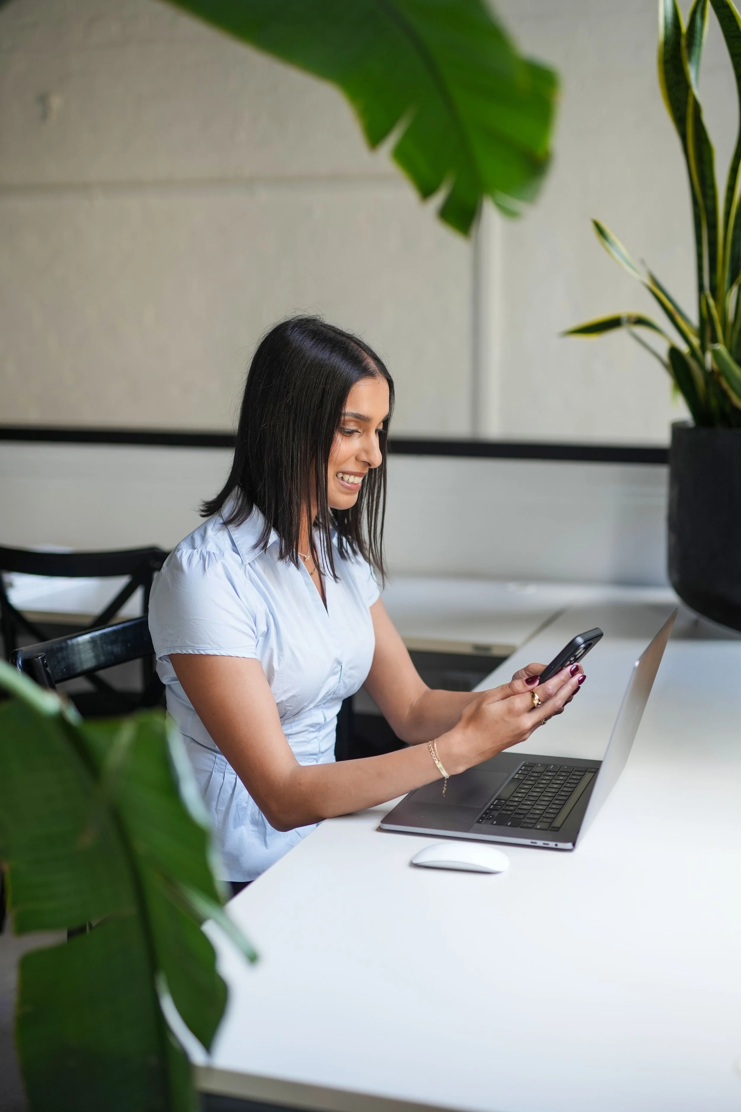 Krysana Chhiba sitting at a white desk, smiling and looking at her phone, with a laptop, a computer mouse, and large green houseplants nearby.