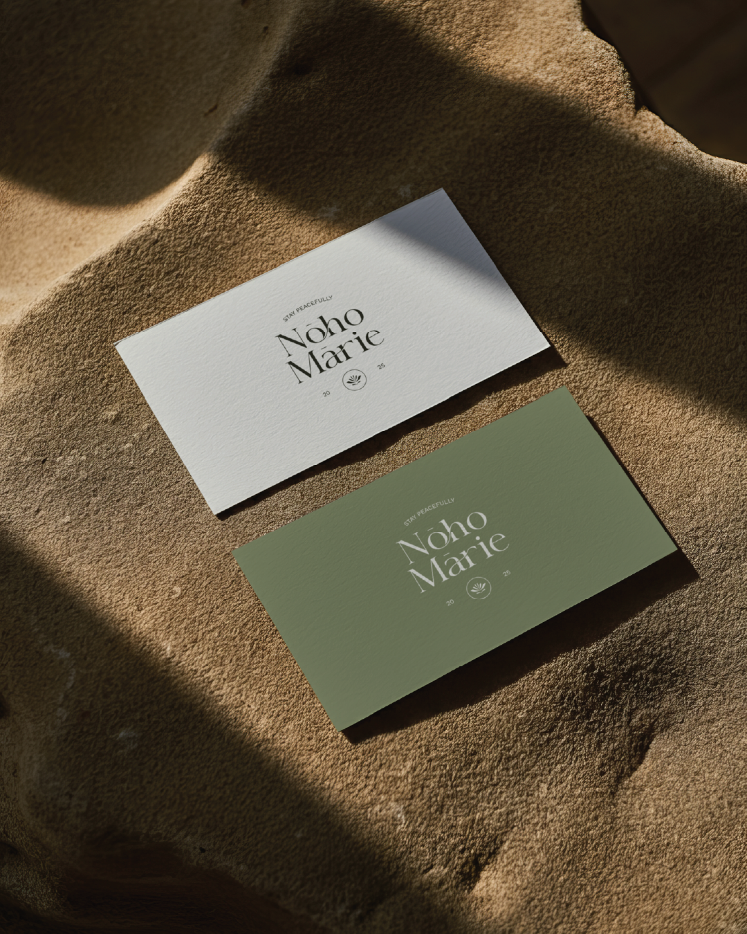 Two business cards for Noho Marie, one white and one green, placed on a textured surface with shadows.