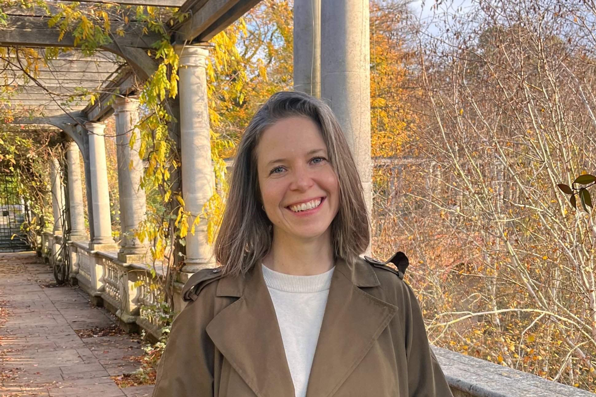 A young woman smiling outdoors in autumn, wearing a tan jacket, standing on a brick path with autumn-colored trees and ornate stone columns in the background.