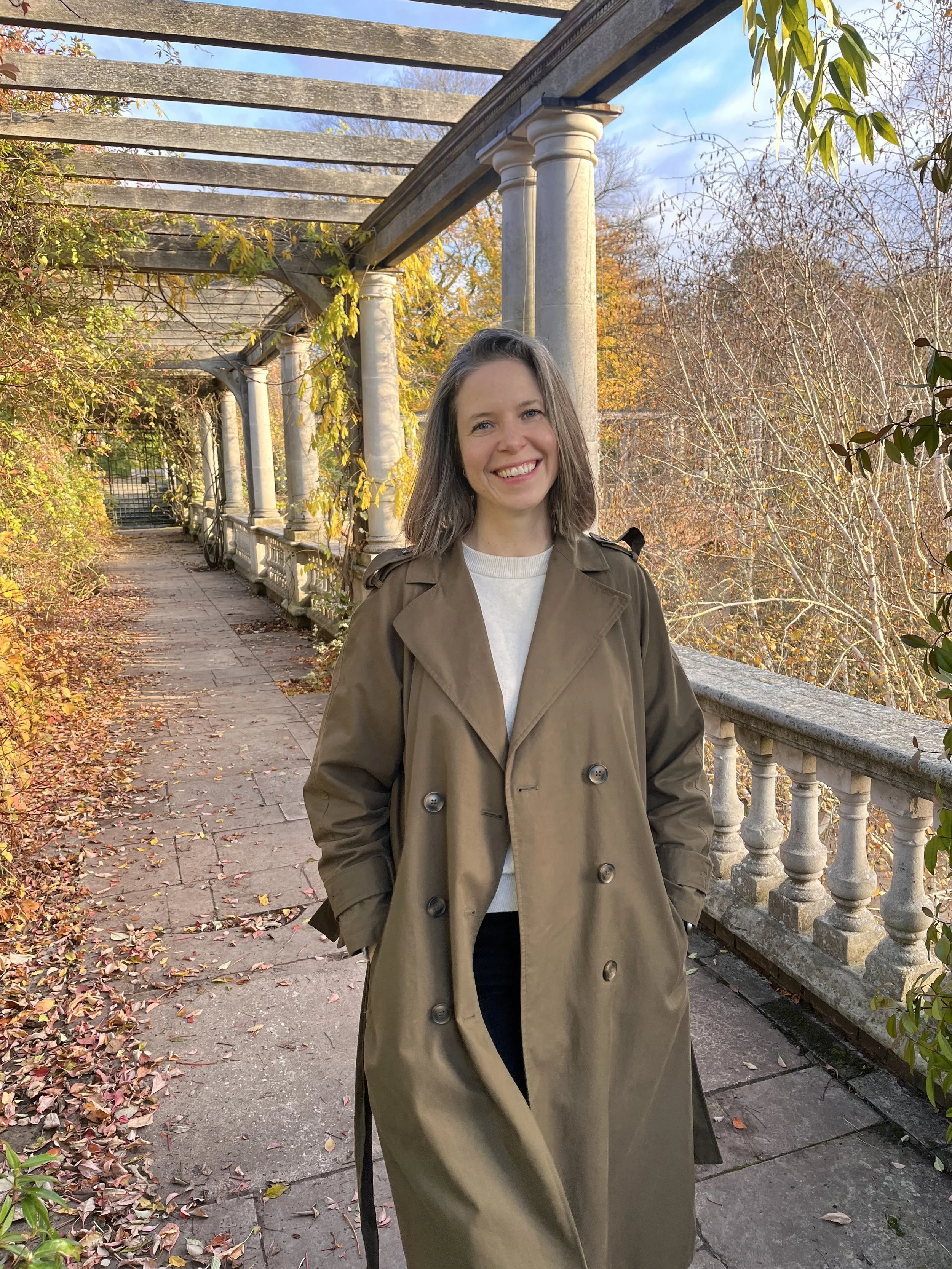 A woman with shoulder-length hair, smiling and wearing a tan trench coat, standing on a pathway with autumn leaves. The background features a pergola with columns and a railing, surrounded by fall foliage.