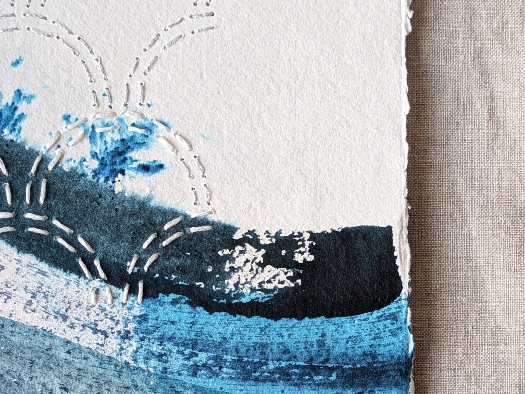 Close-up of hand sewn textile details on handmade indian khadi paper art piece featuring embroidered white dotted lines and abstract blue washes with torn edges and a background of brown fabric.