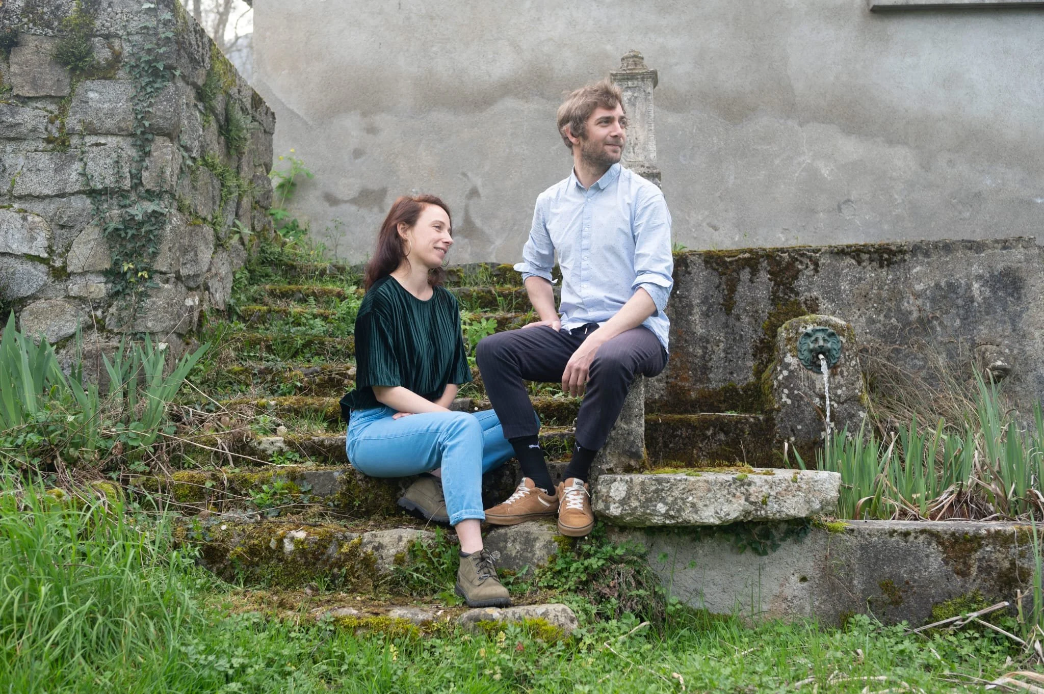 Deux personnes assises sur des escaliers en pierre dans un jardin avec une fontaine en pierre, un homme et une femme, entourés de végétation, souriant et se détendant.