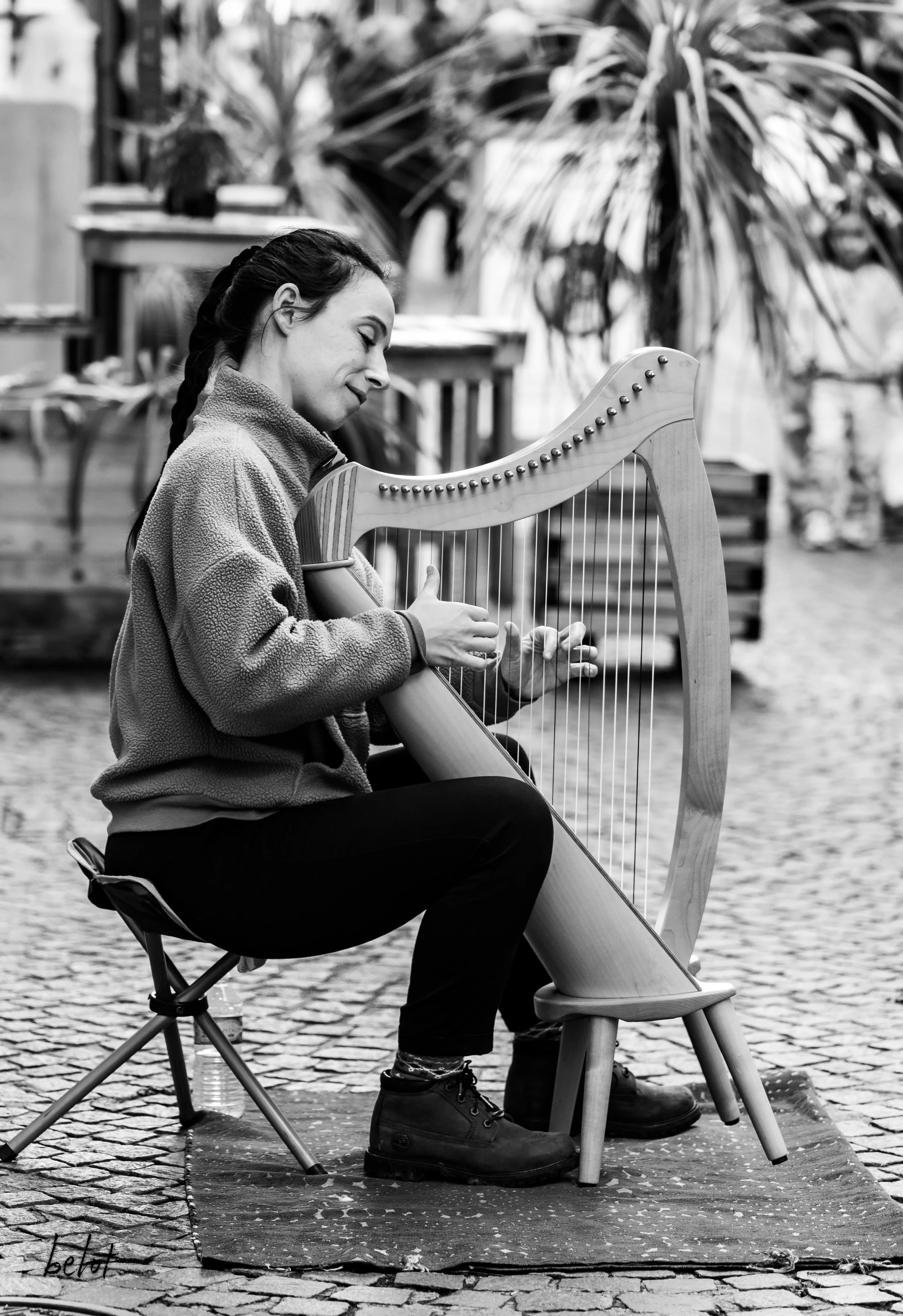 Une femme jouant de la harpe assise sur une chaise, en extérieur, avec des plantes en arrière-plan. Photo en noir et blanc.